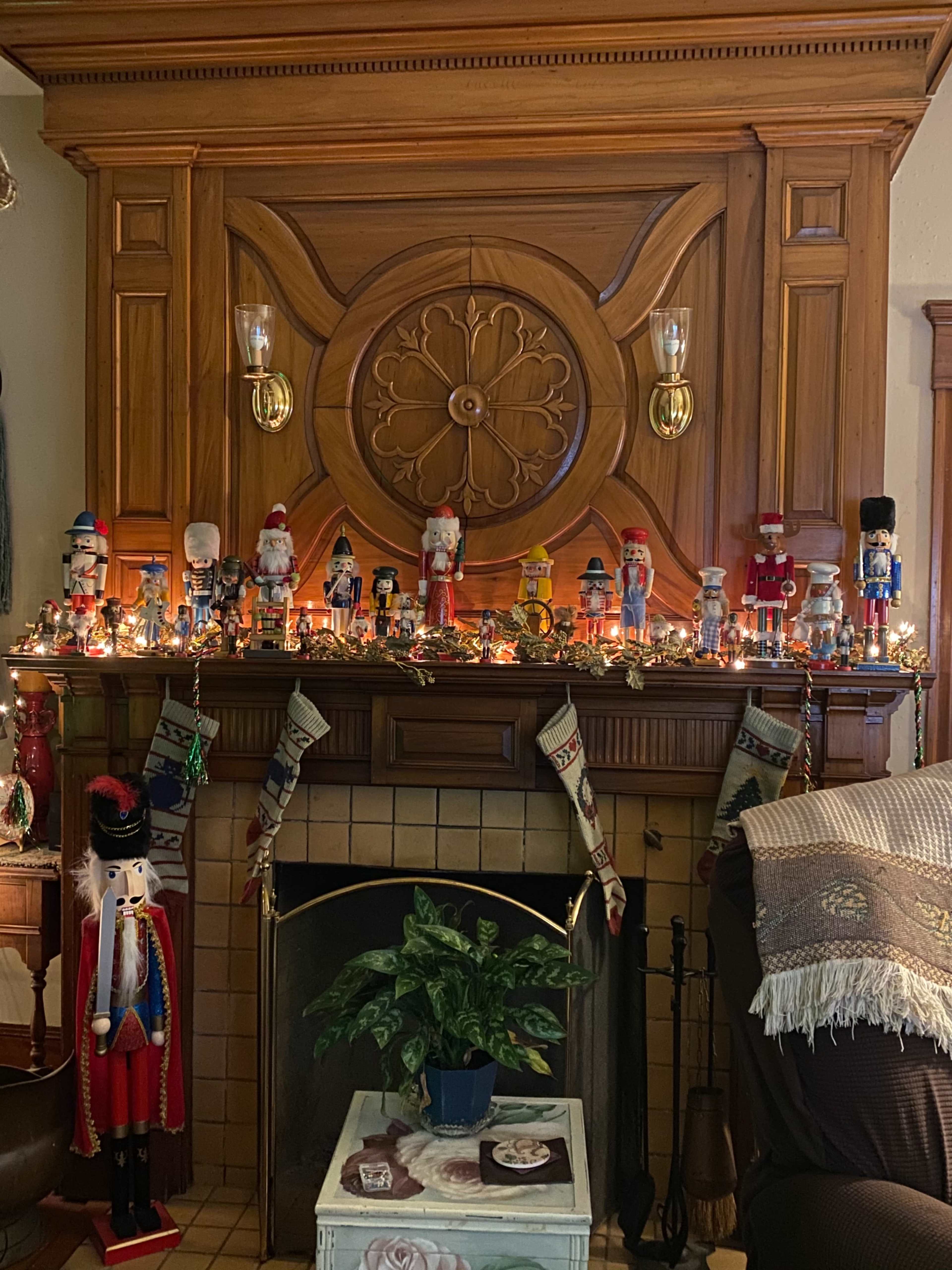 A wooden mantel decorated with a festive arrangement of nutcracker figurines, twinkling lights, and Christmas stockings, with a plant and a cozy armchair nearby.
