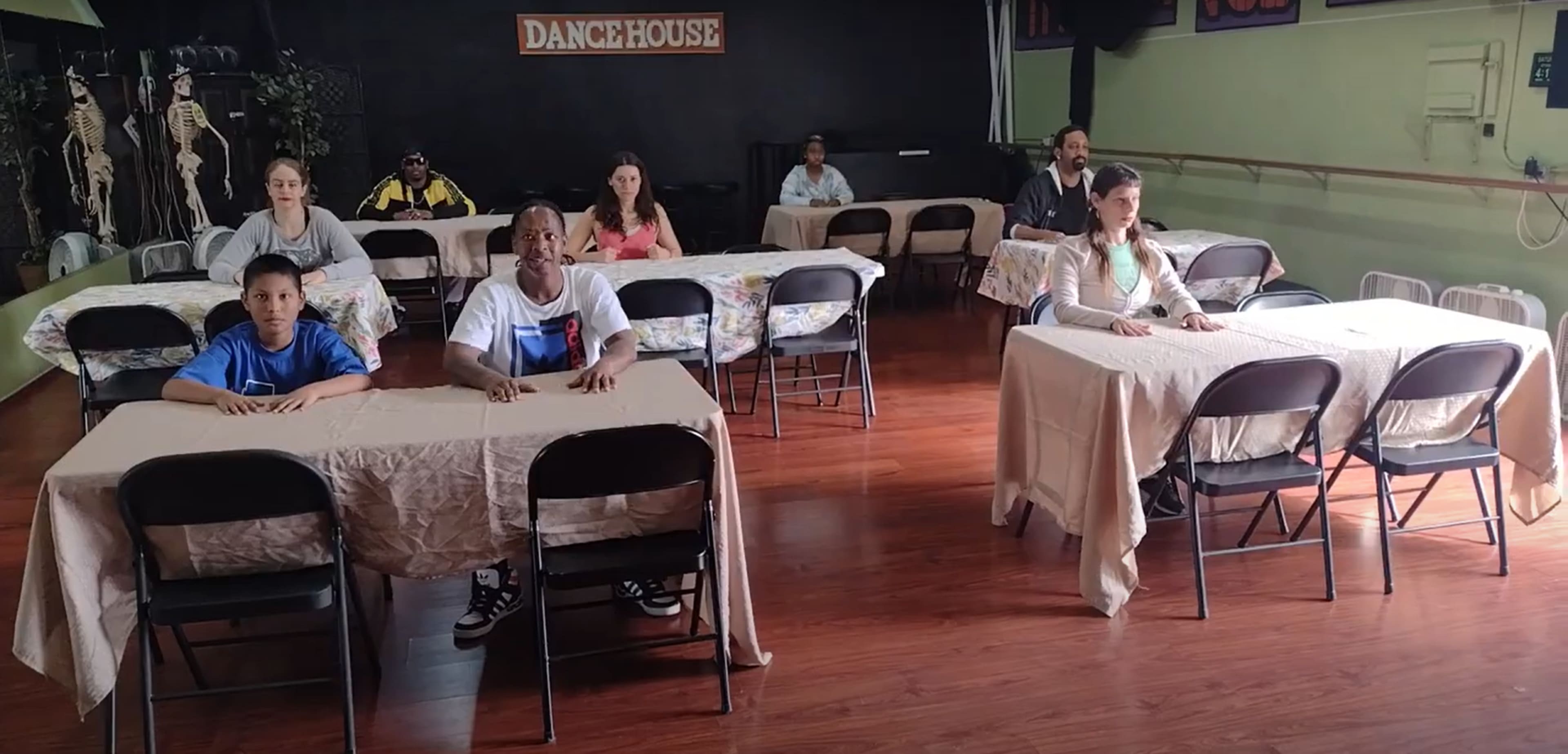 A group of people sits at tables in a dance studio, facing forward with a casual arrangement of chairs and tablecloths.