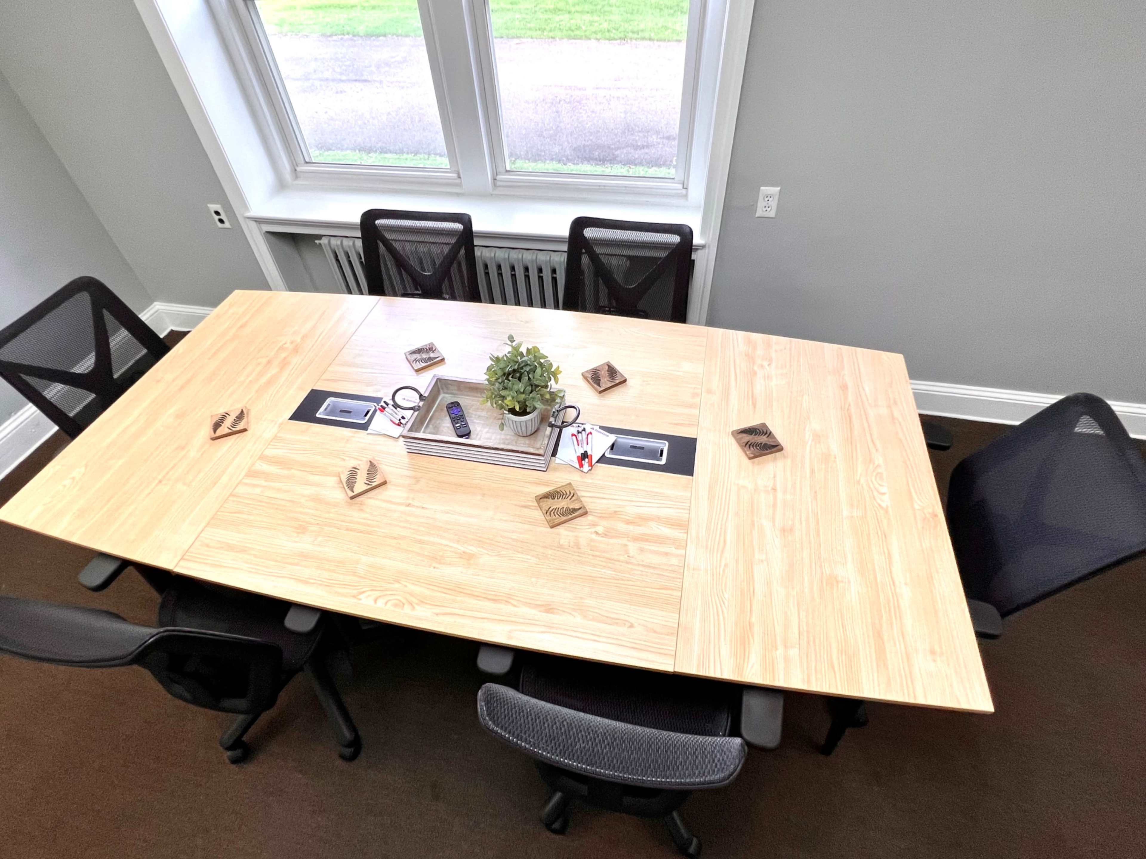 A large wooden conference table is set in a well-lit room, surrounded by several black office chairs, with a small plant and office supplies on the table.