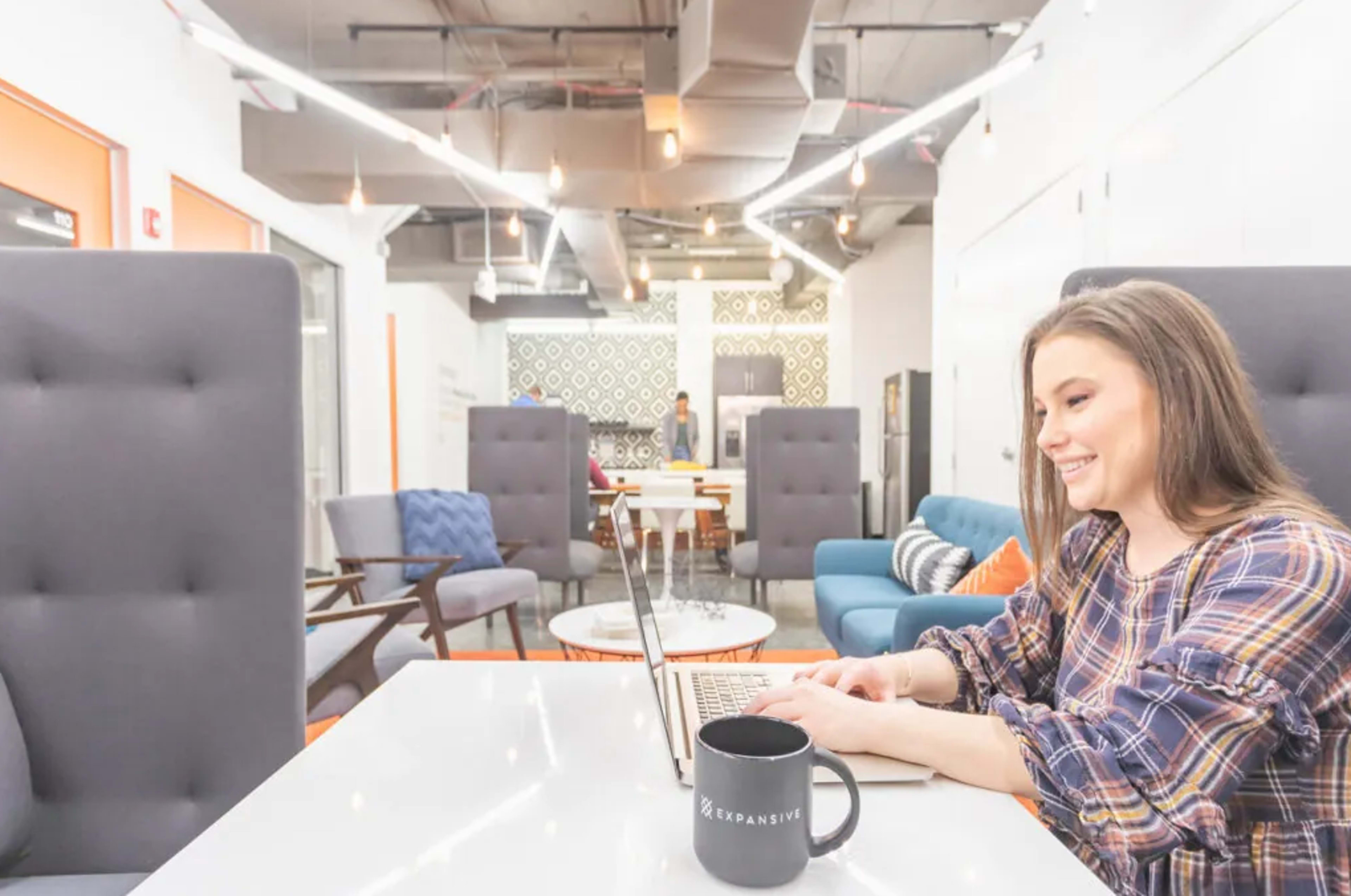 A woman is working on a laptop at a table in a modern office space with colorful furniture and a bright atmosphere.
