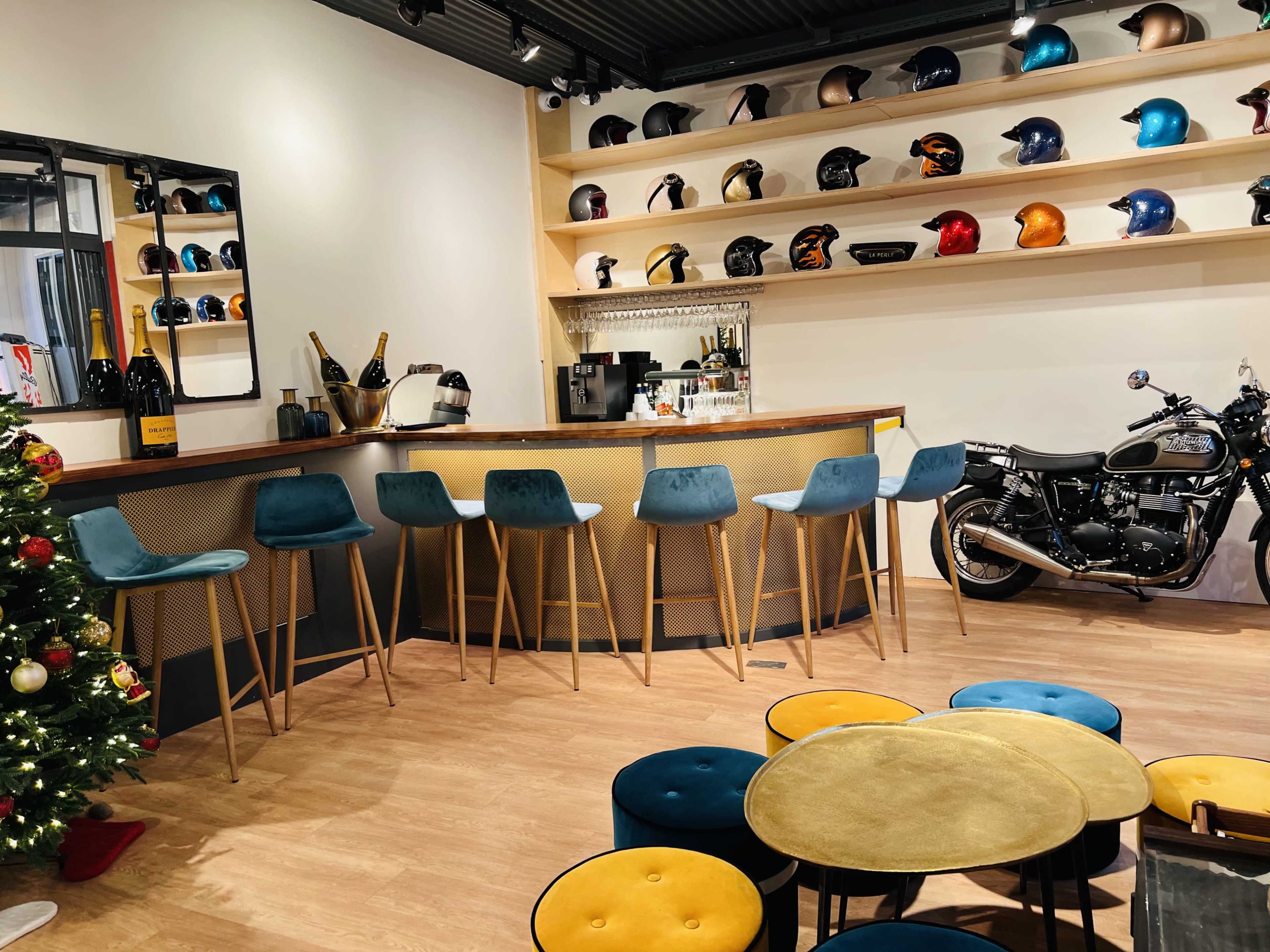 The image depicts a stylish bar area with blue stools, a motorcycle displayed on the right, and shelves lined with various motorcycle helmets.