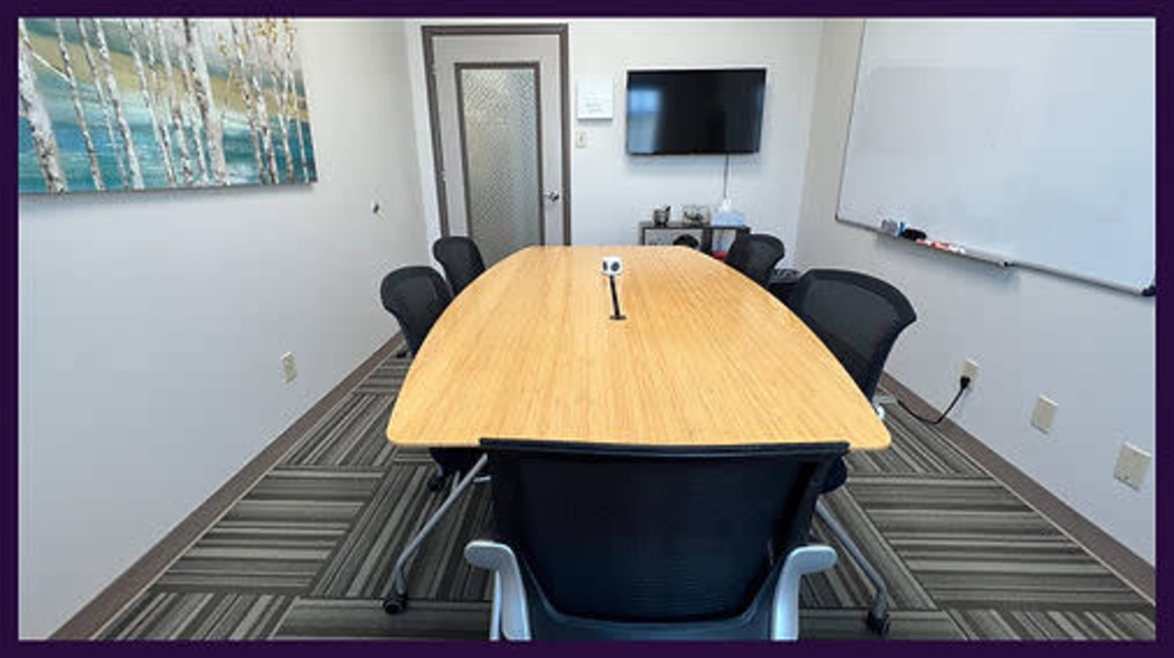 A modern conference room features a long wooden table surrounded by black chairs, with a wall-mounted TV and whiteboard in the background.