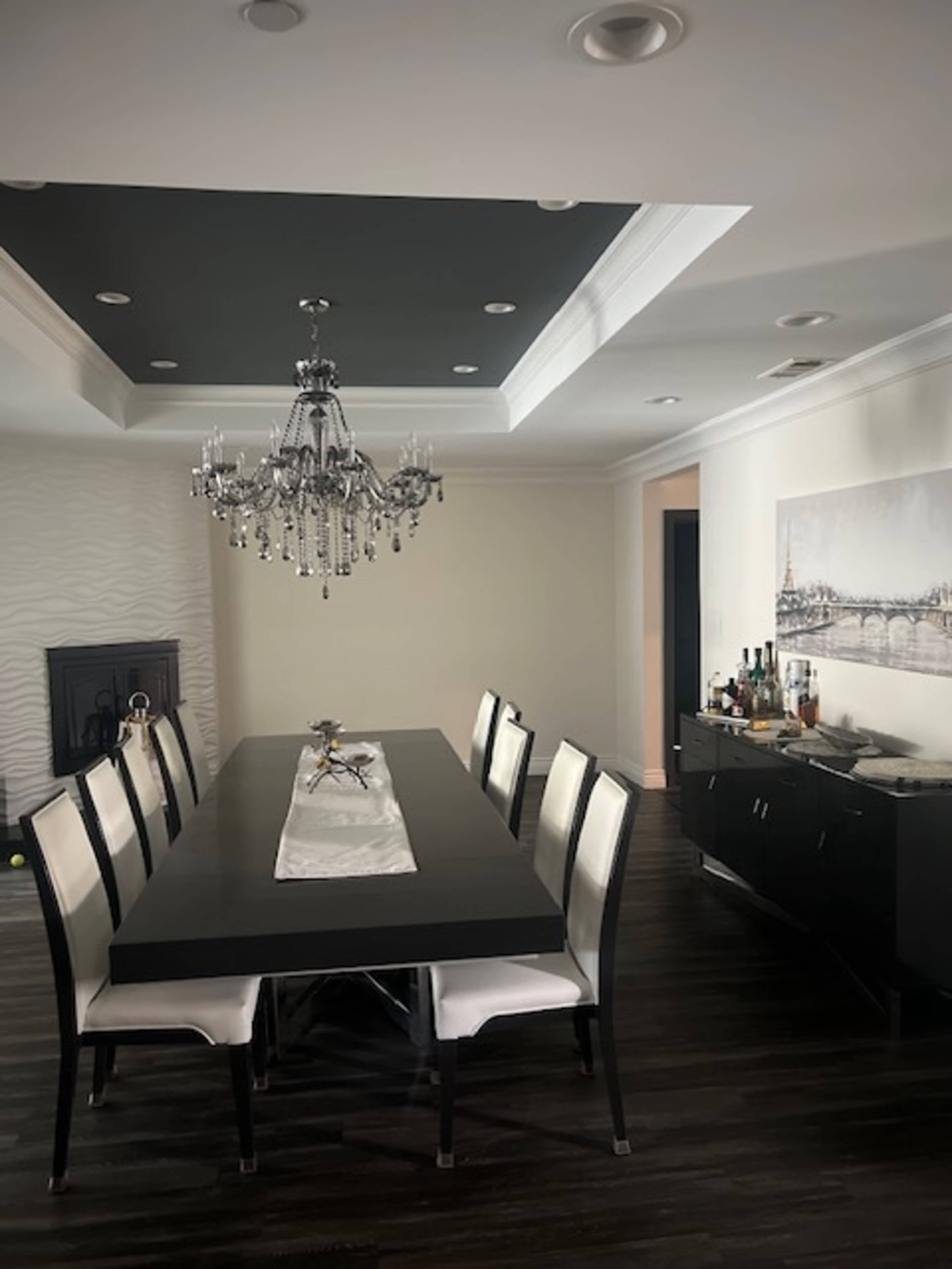 The image shows a modern dining room featuring an elaborate chandelier above a long black table surrounded by white and black chairs, with a sideboard displaying various beverages.
