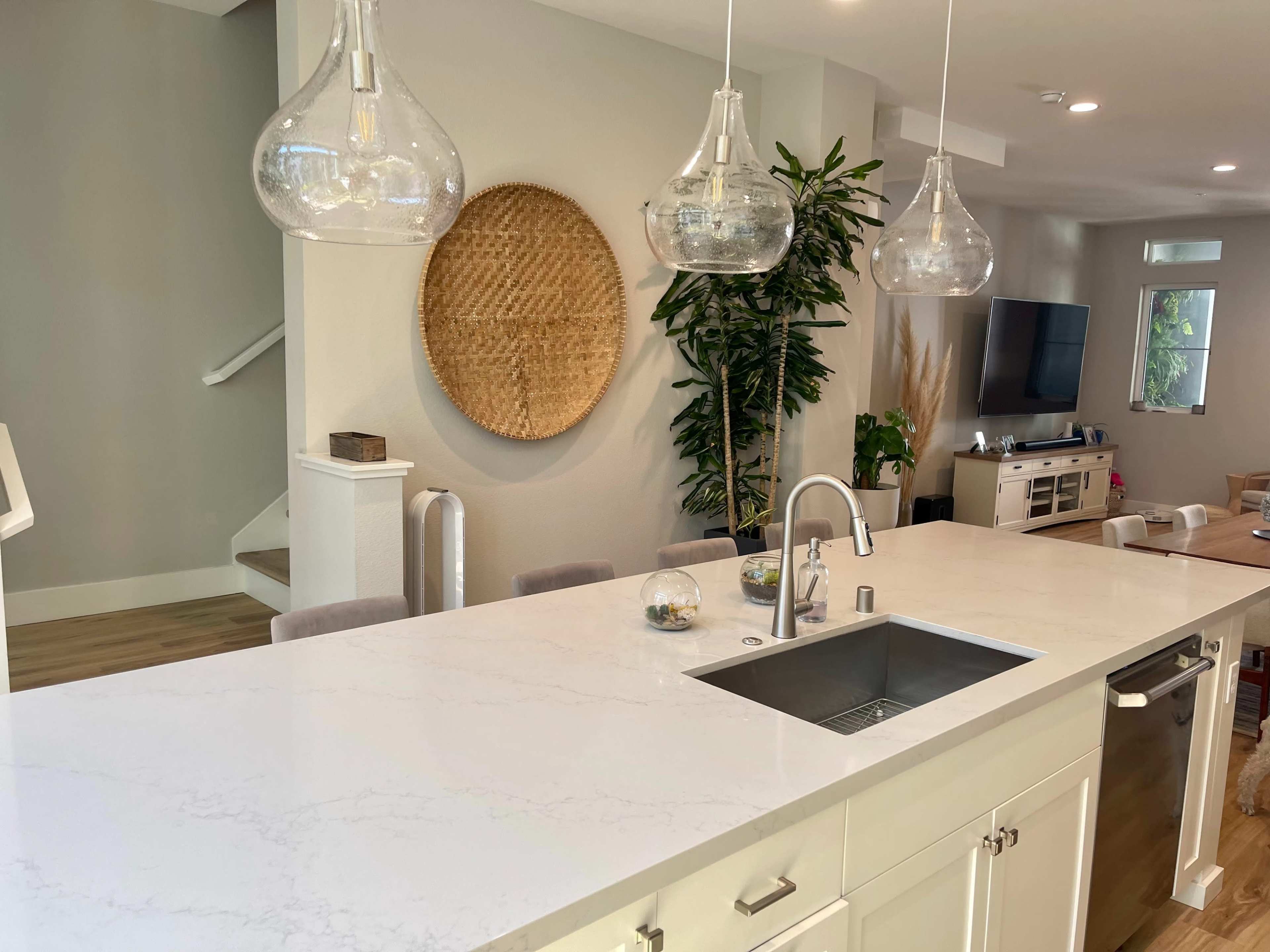 A modern kitchen with a large white countertop, pendant lights, a stainless steel sink, and a decorative woven wall piece.