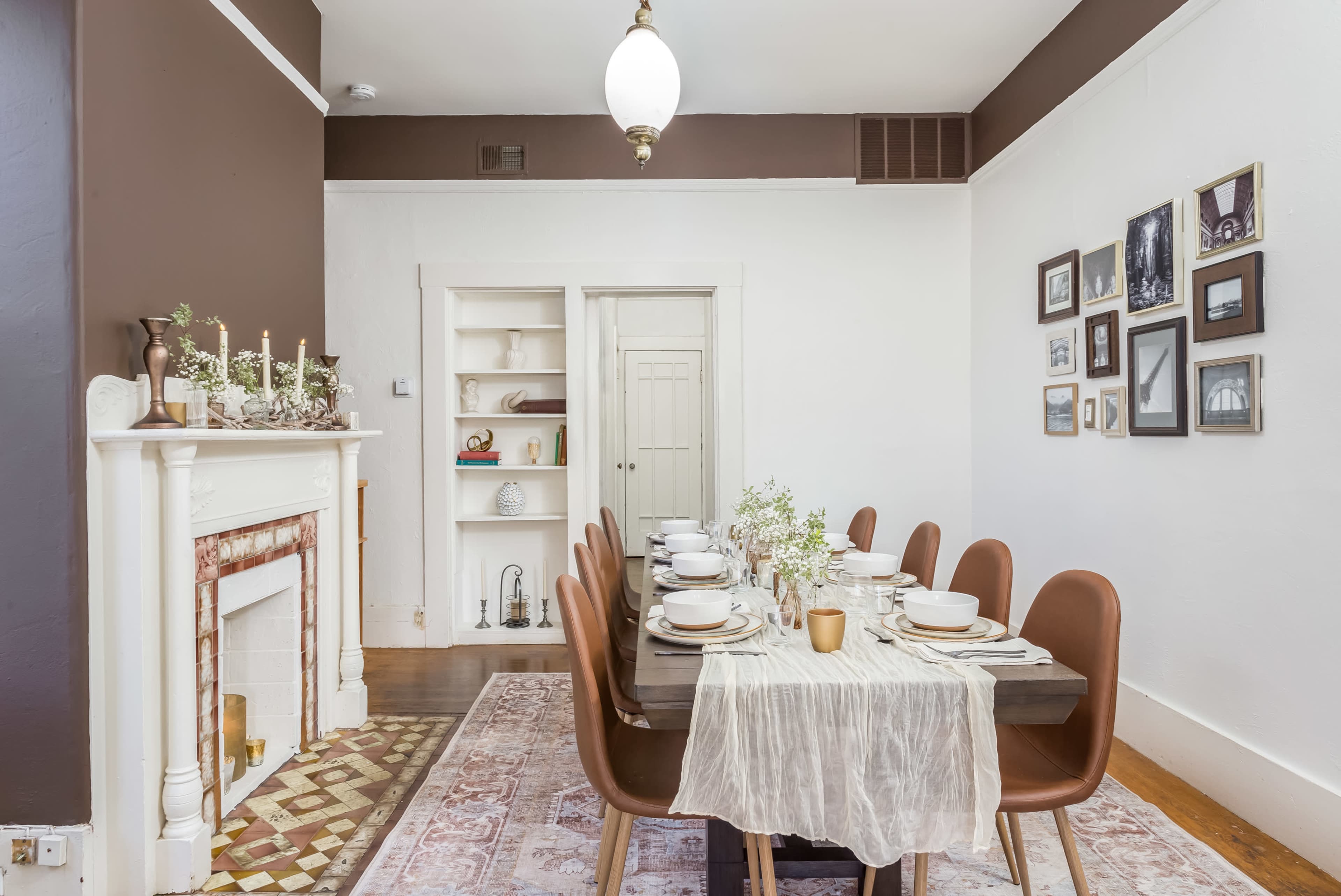 A long dining table is set with plates and utensils in a room with a white fireplace, brown walls, and framed pictures on the adjacent wall.