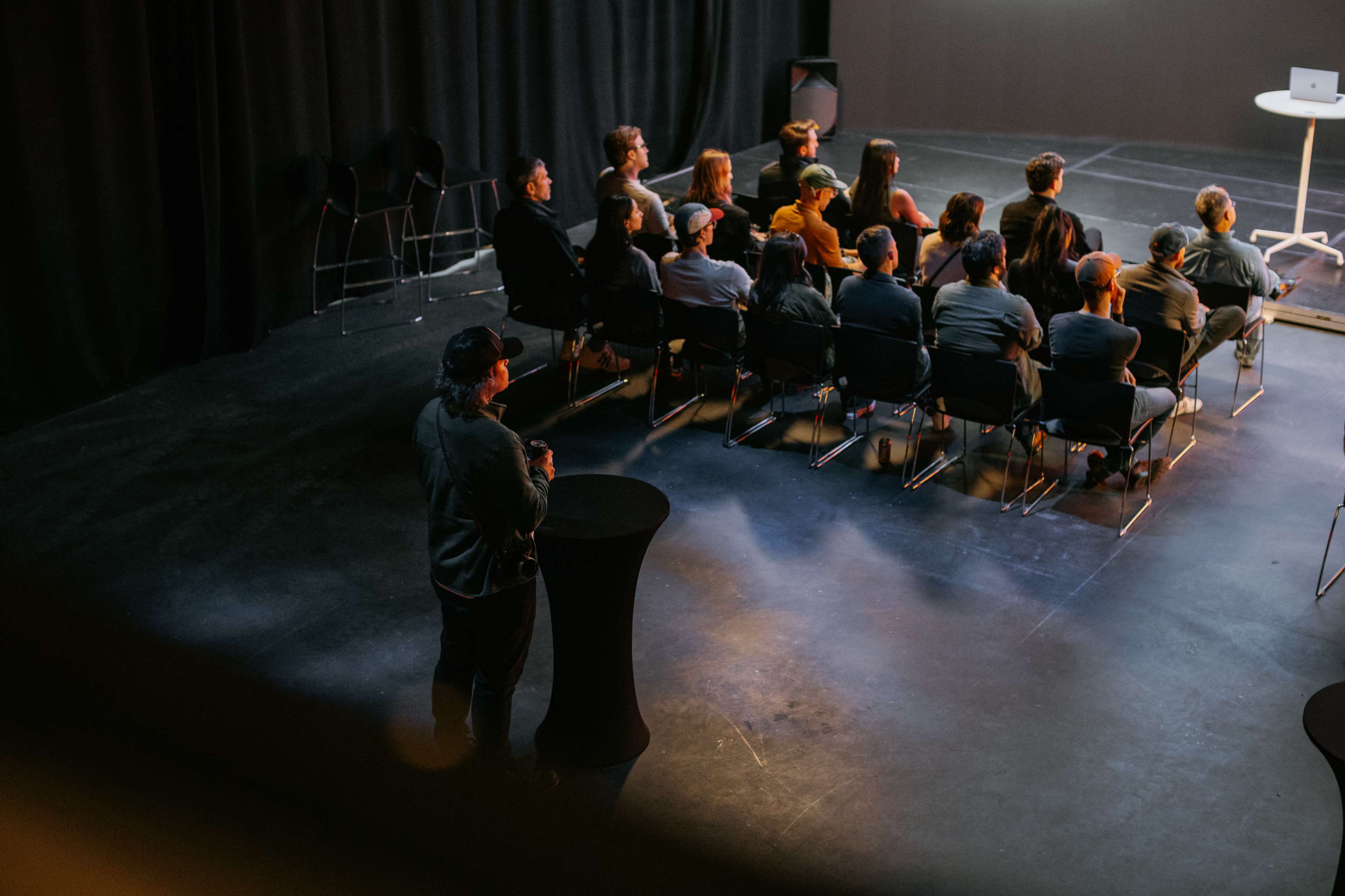 A group of people sits in chairs facing a presentation area, while another person stands near a high table at the side of the room.