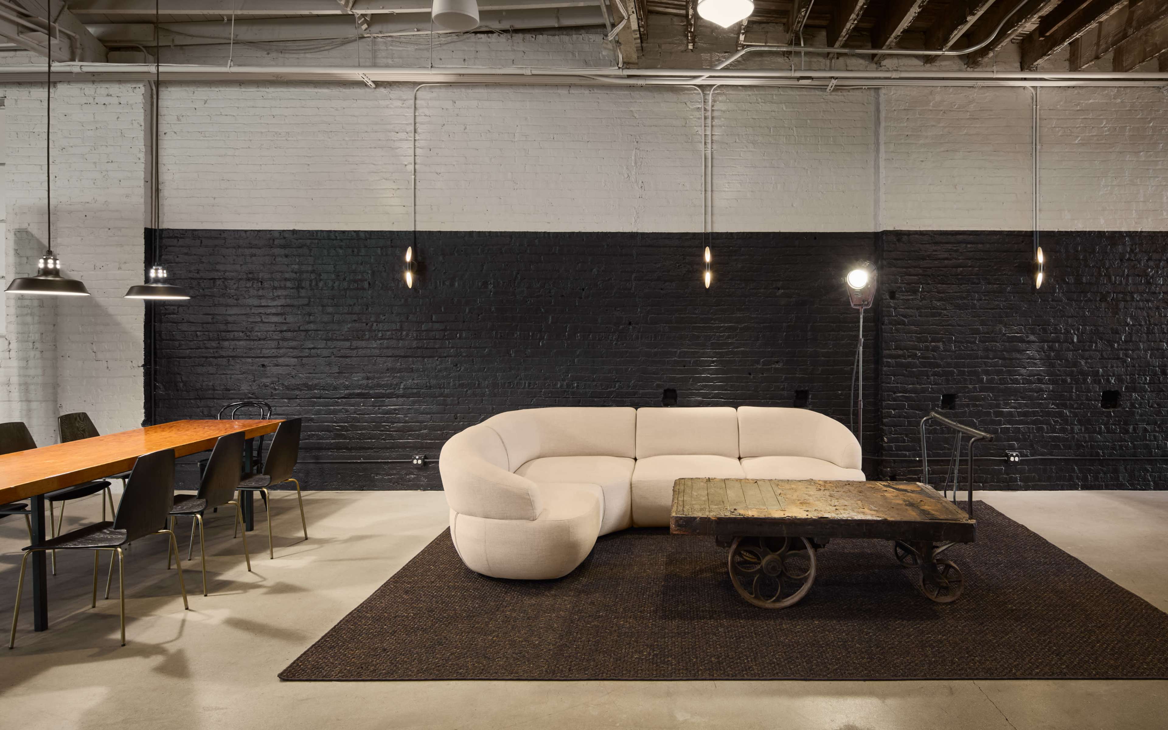 The image depicts a modern interior with a light-colored curved sofa, a rustic wooden table on a dark area rug, and a long dining table in a room with exposed brick walls and industrial lighting.