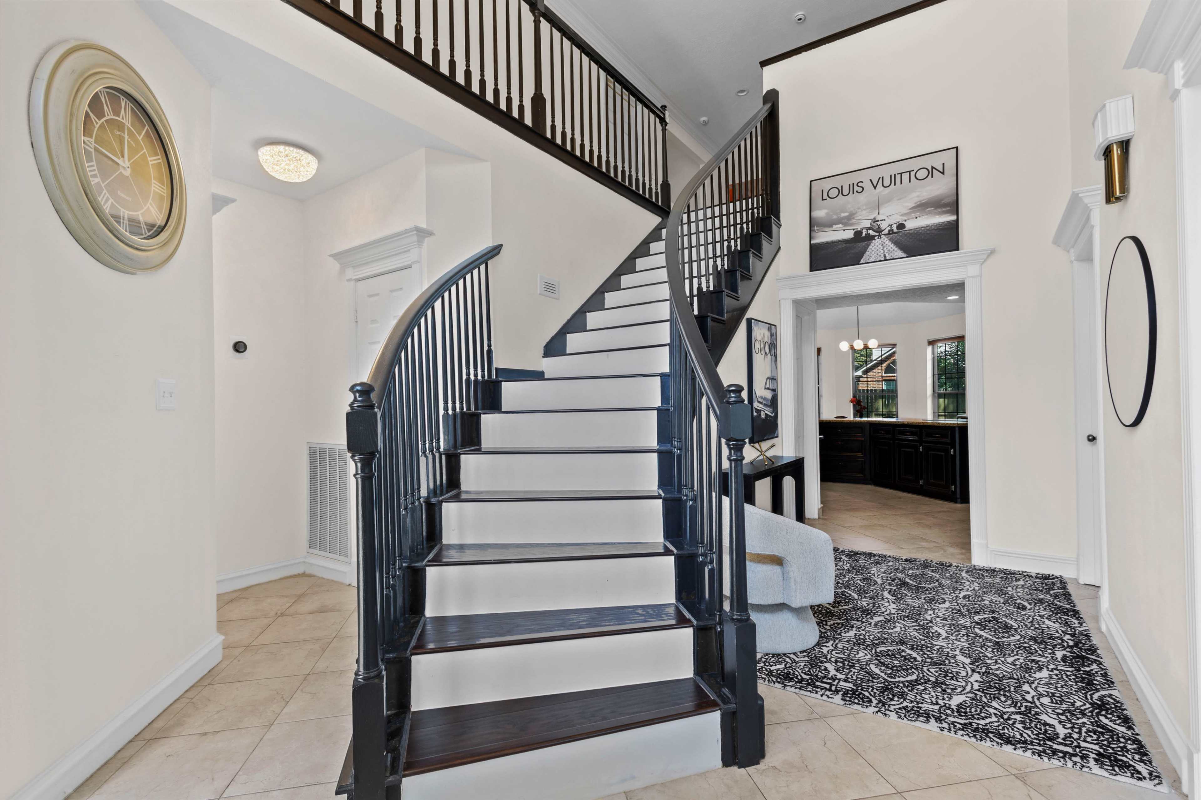 A curved staircase with black railings is the focal point of a spacious entryway featuring tile flooring and a wall adorned with a Louis Vuitton print.
