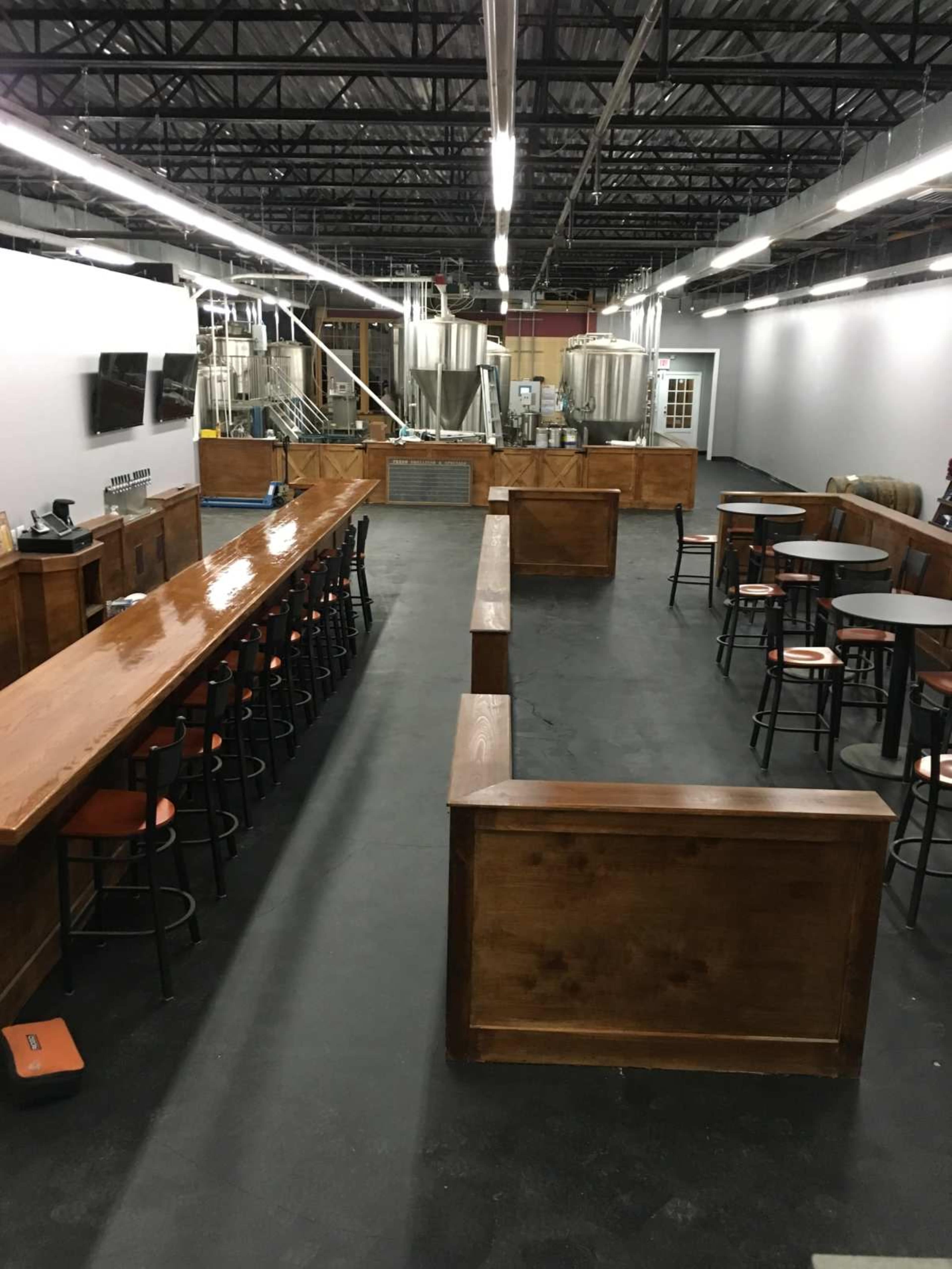 The image shows a spacious brewery interior with wooden counters, bar stools, and stainless steel tanks in the background.