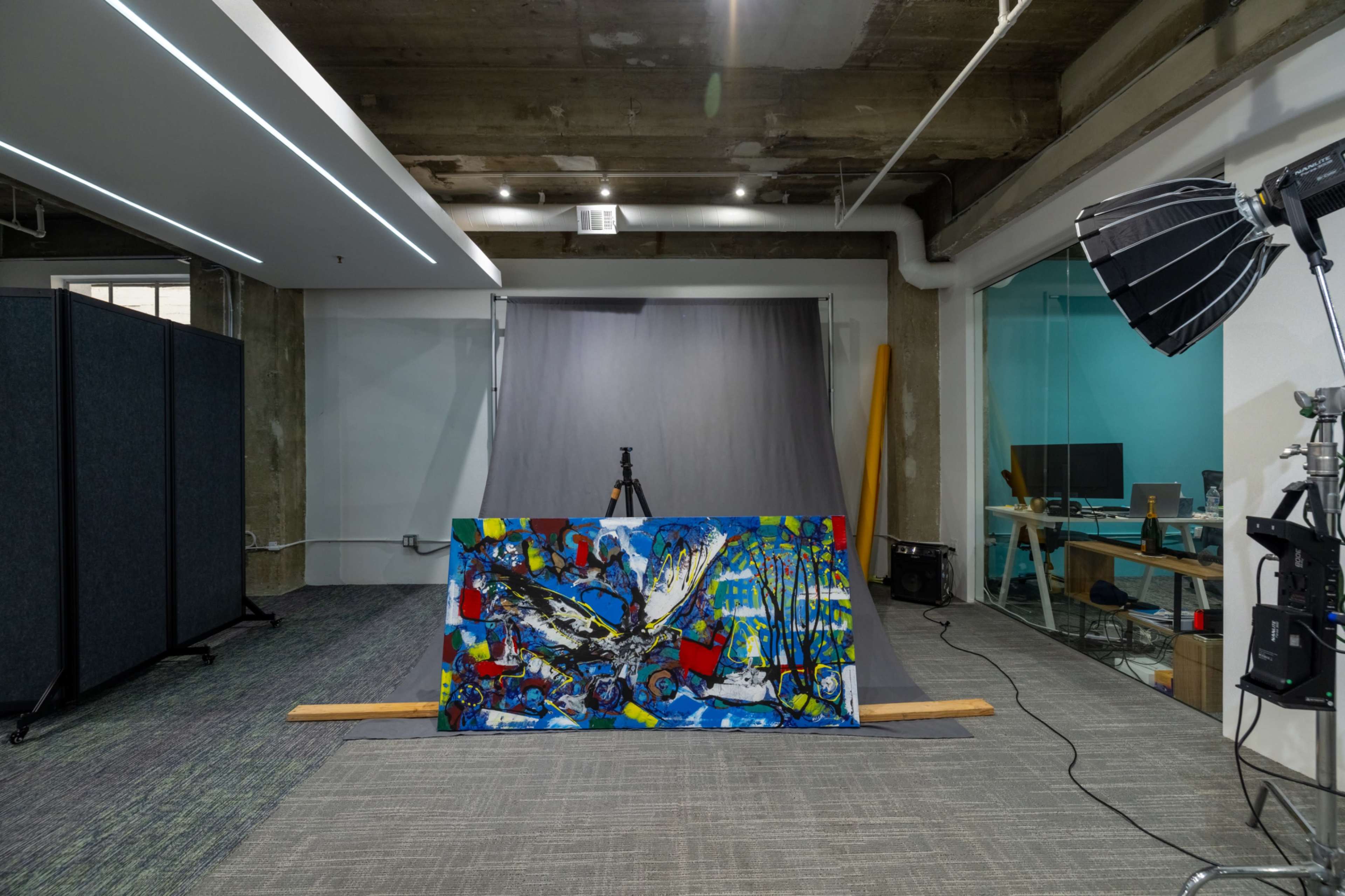 The image shows an art studio with a colorful painting on display, positioned in front of a photography setup, including lights and a backdrop.