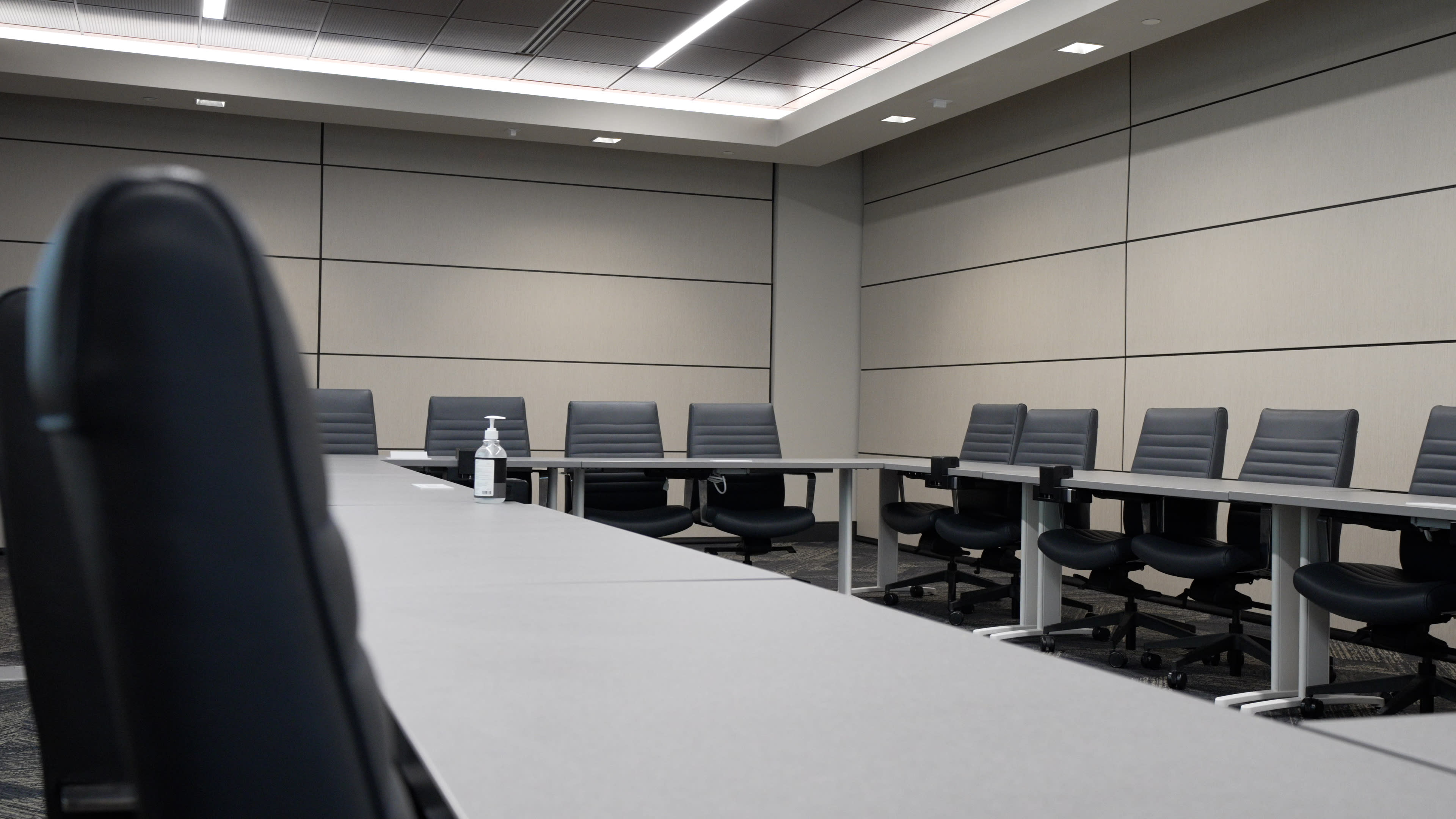 A large conference room features a long table surrounded by multiple black chairs, with a lone water bottle placed on the table.