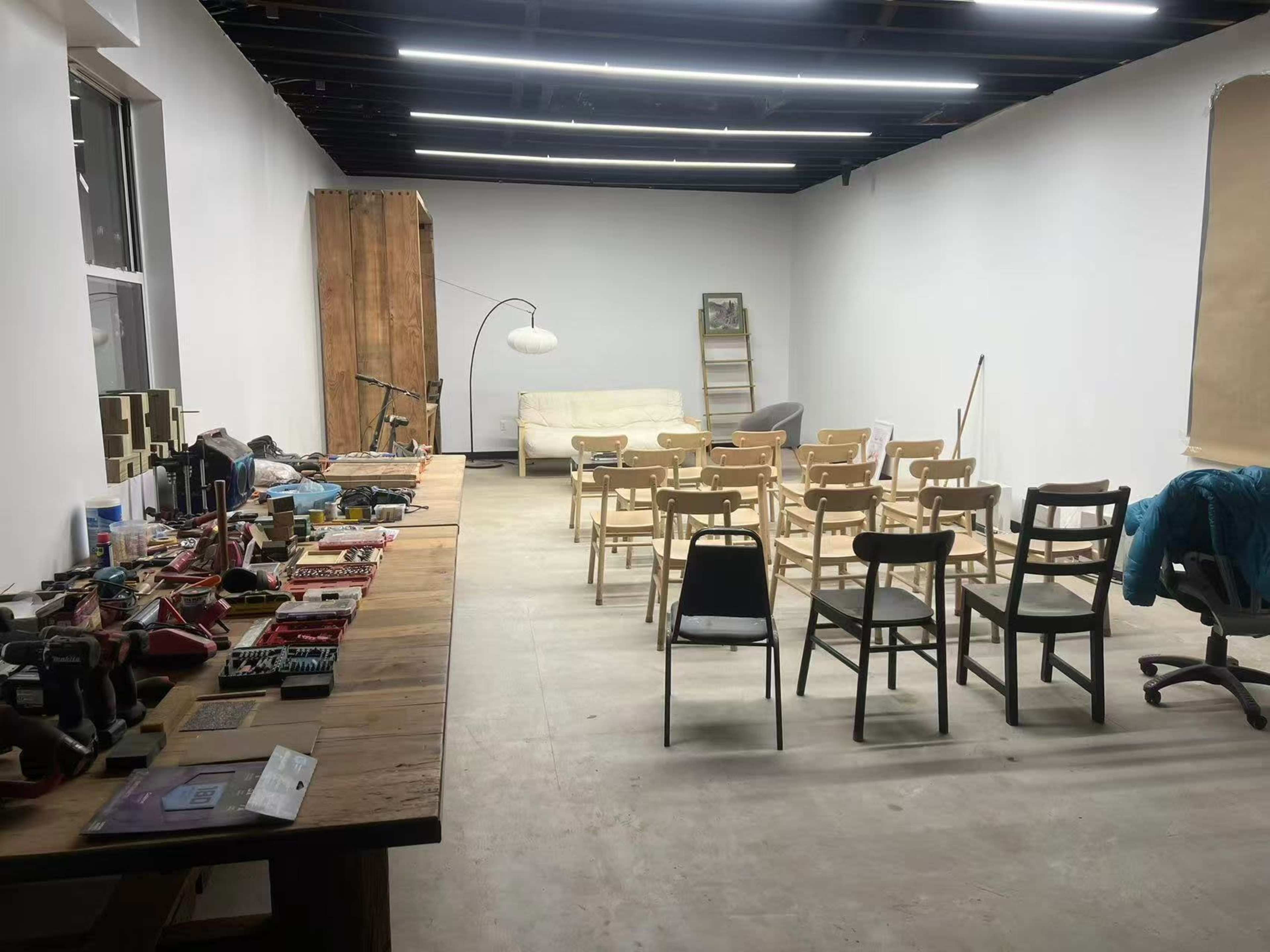 A spacious, unfinished room features a wooden table with various tools and supplies on one side and rows of wooden chairs facing a blank wall on the other.