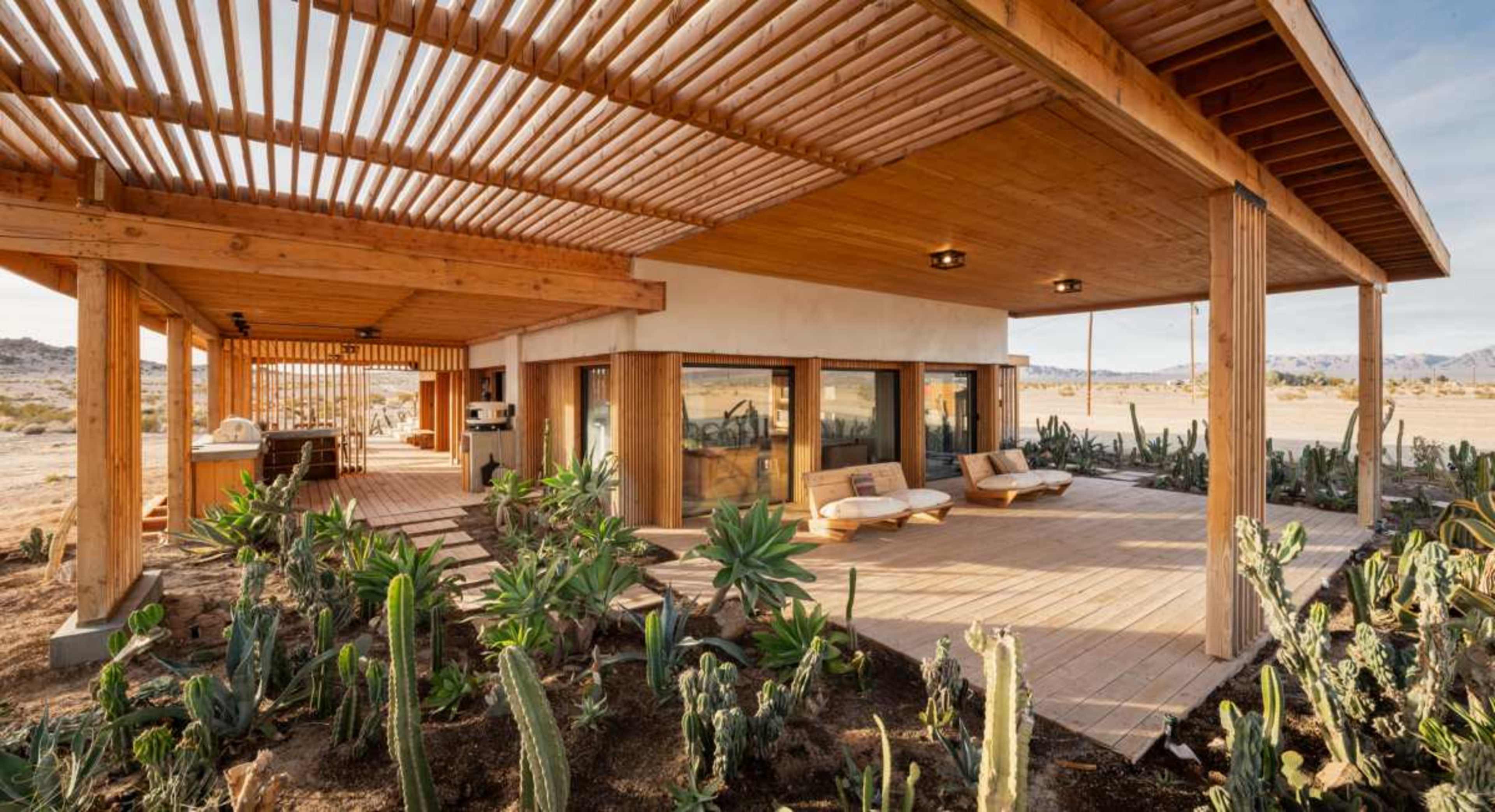 The image shows a modern single-story house with a wooden overhang, large windows, and a patio surrounded by desert vegetation.