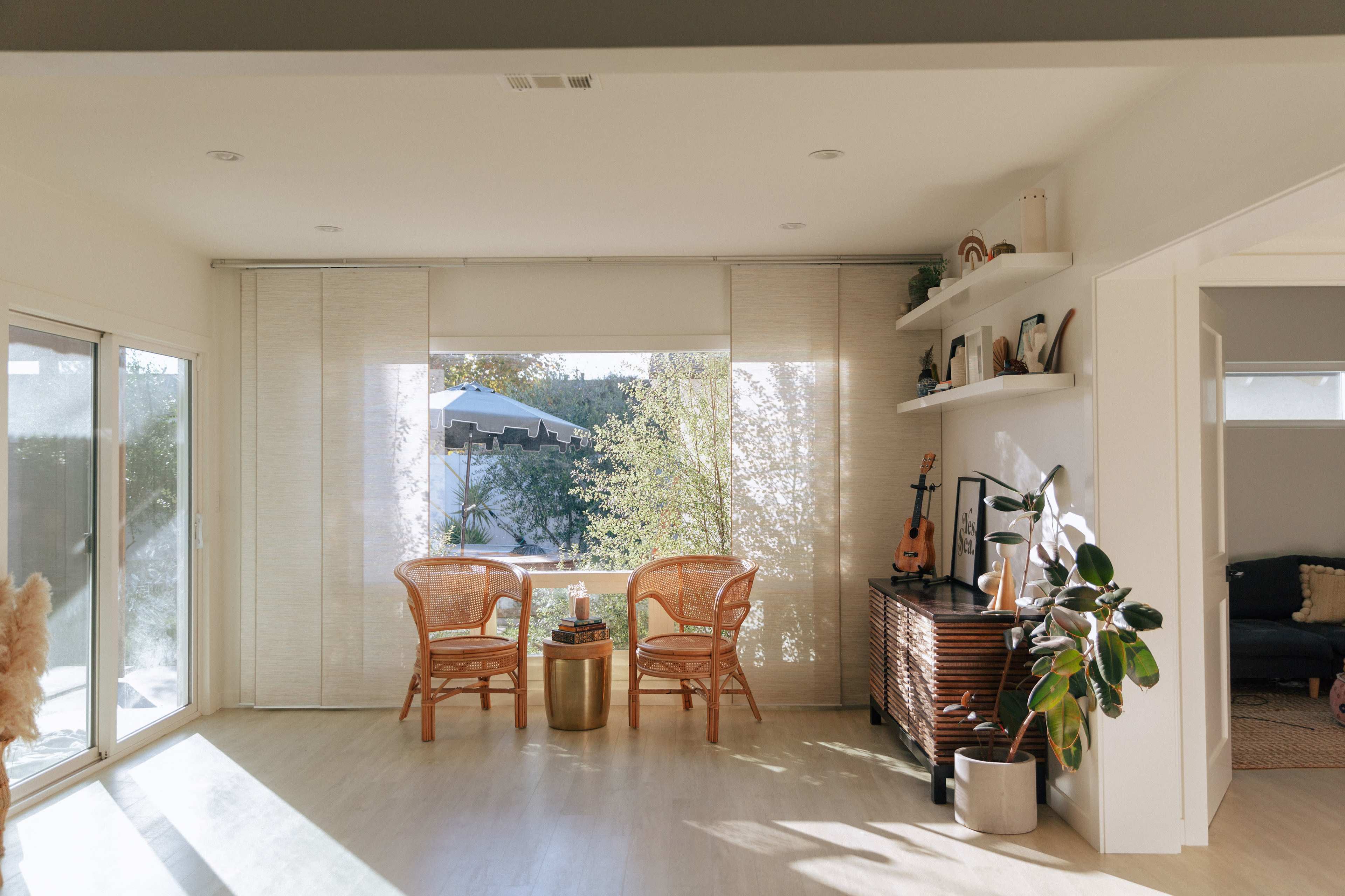 A bright living room features two rattan chairs facing a large window, with plants and decor on a shelf and a media console nearby.