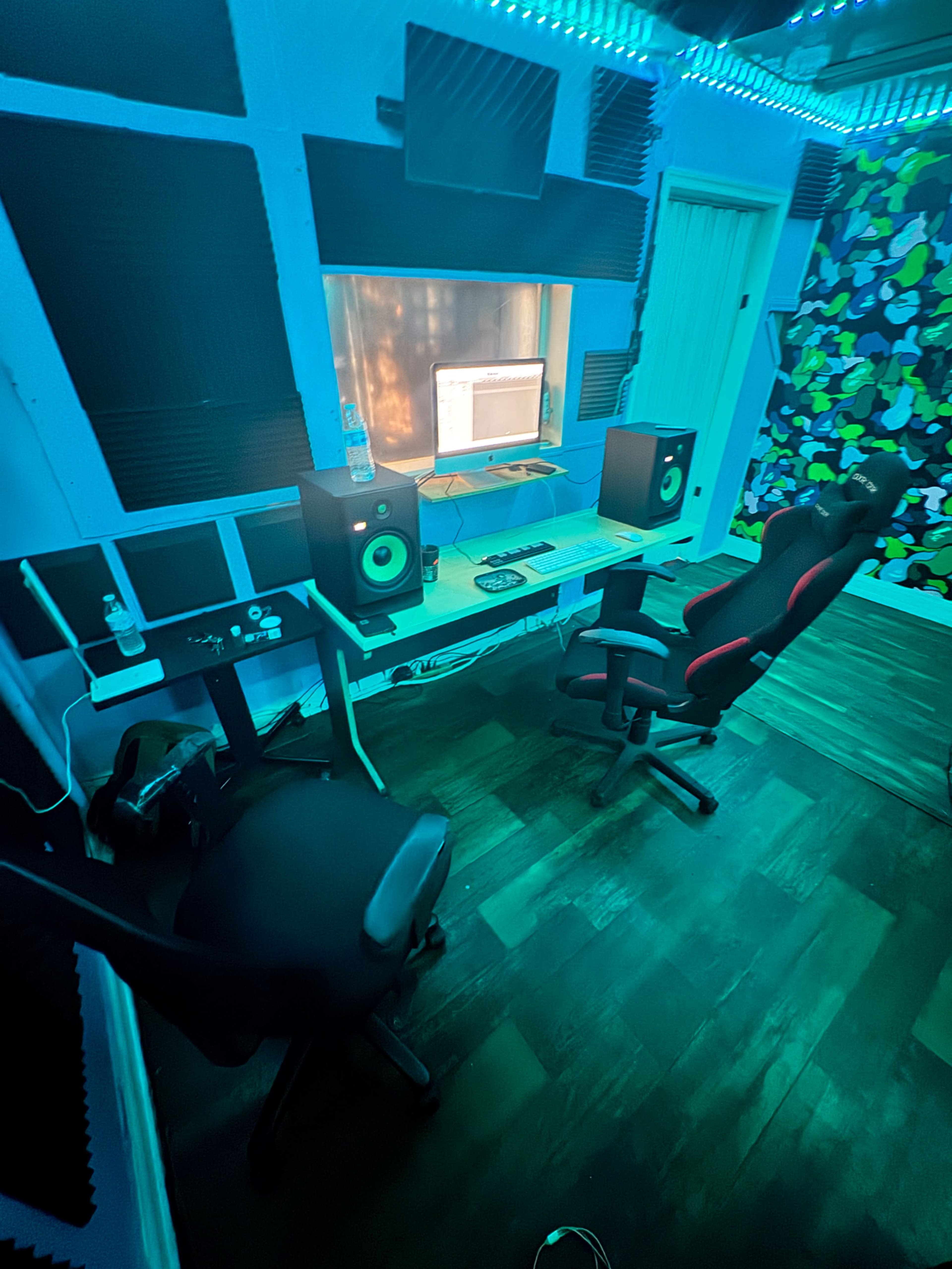 The image shows a small music studio with a desk, two chairs, and monitors, all illuminated by blue and green LED lights.