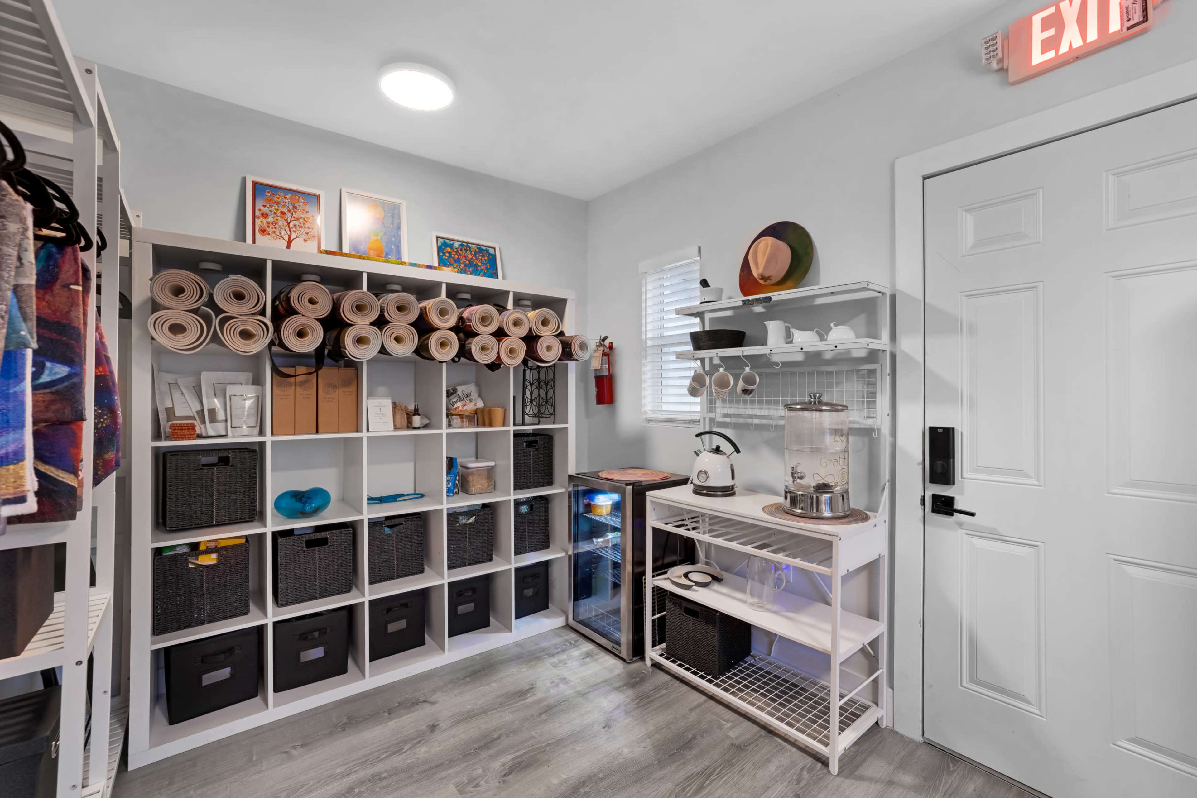The image shows a tidy closet space with organized shelves containing rolls of paper, storage bins, and a small appliance setup, alongside a door marked with an exit sign.