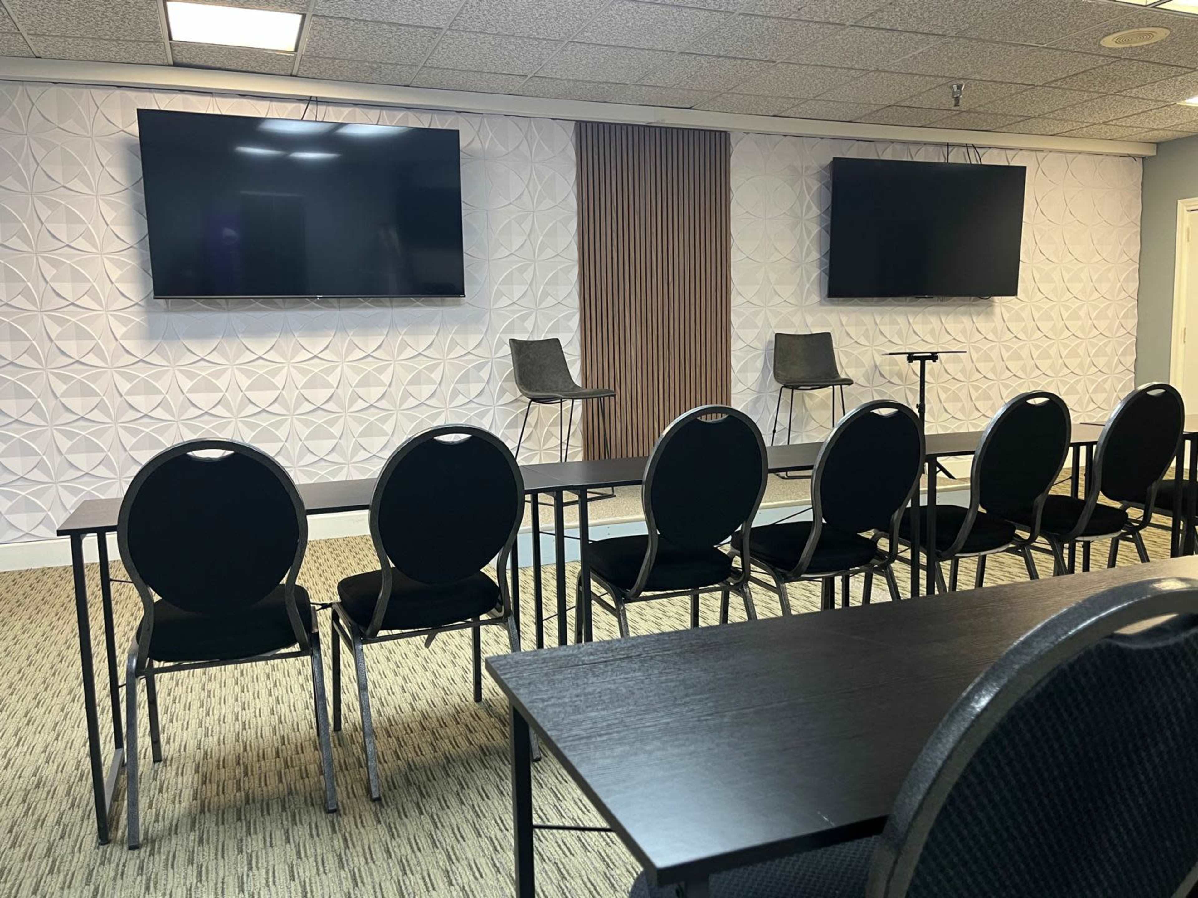The image shows a conference room setup with two large screens, two empty chairs, and rows of black chairs facing a table.