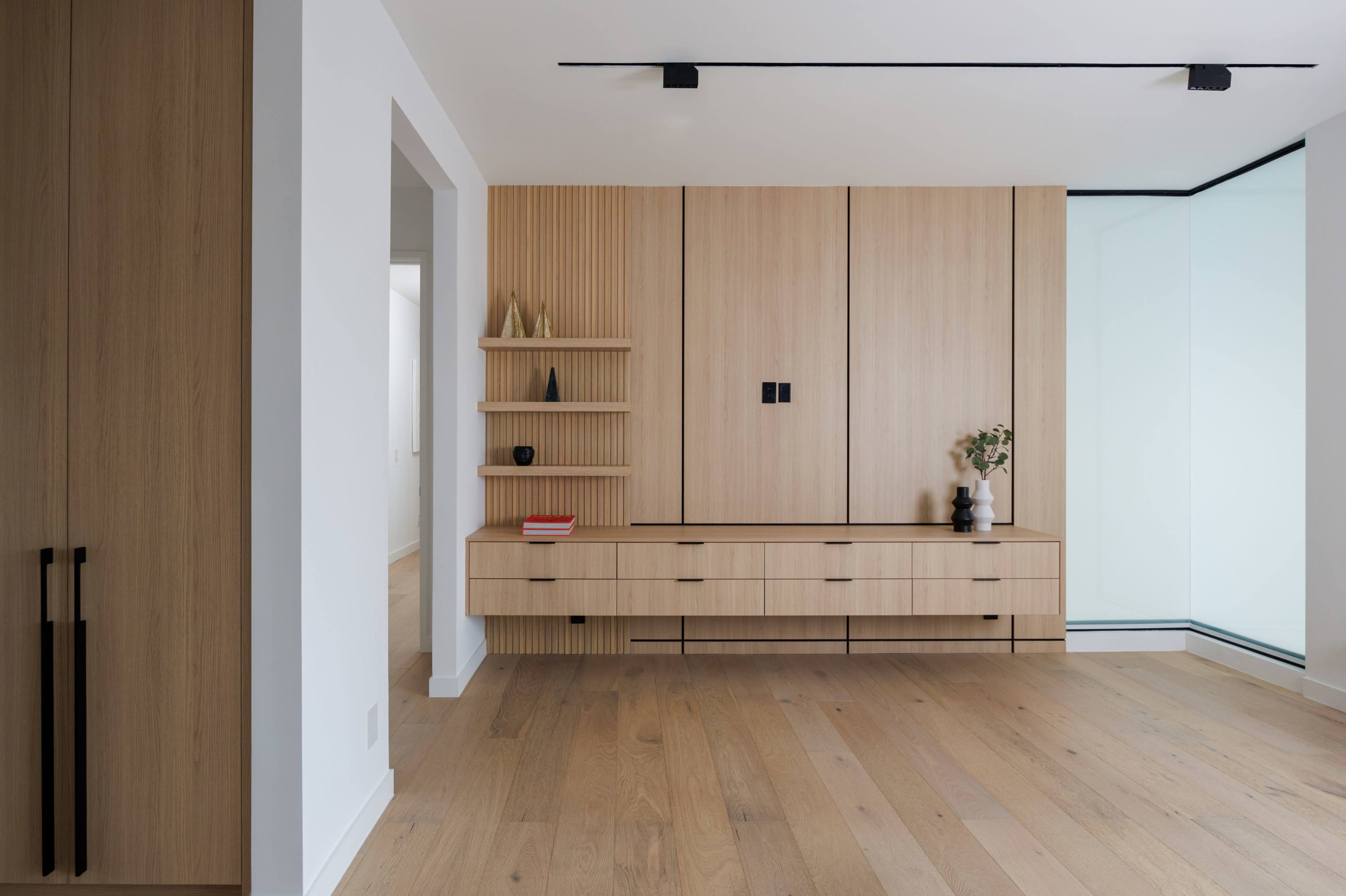The image shows a modern interior with a wooden wall unit featuring drawers, shelves, and a minimalist design, set against a backdrop of light-colored walls and hardwood flooring.