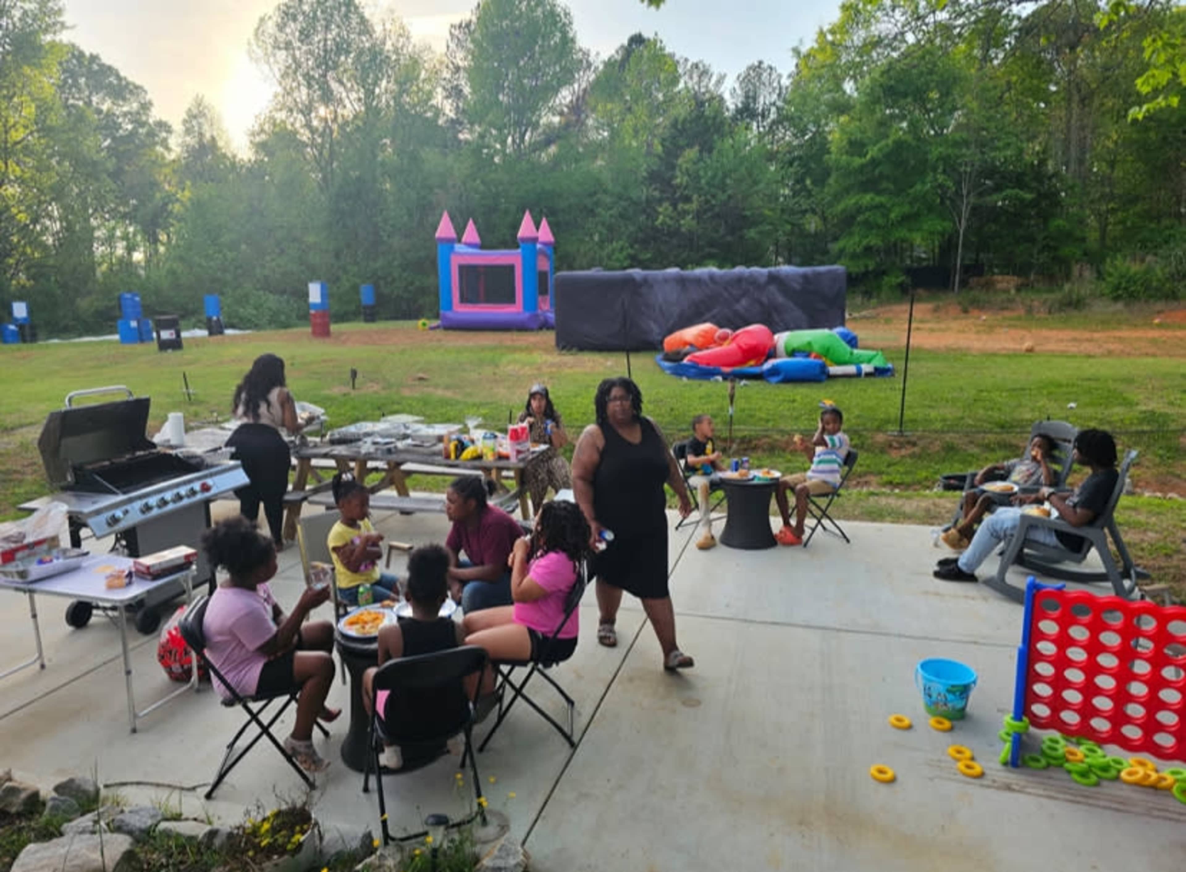 A group of people is gathered at a backyard barbecue, with children playing, a bounce house in the background, and food being served on a patio.