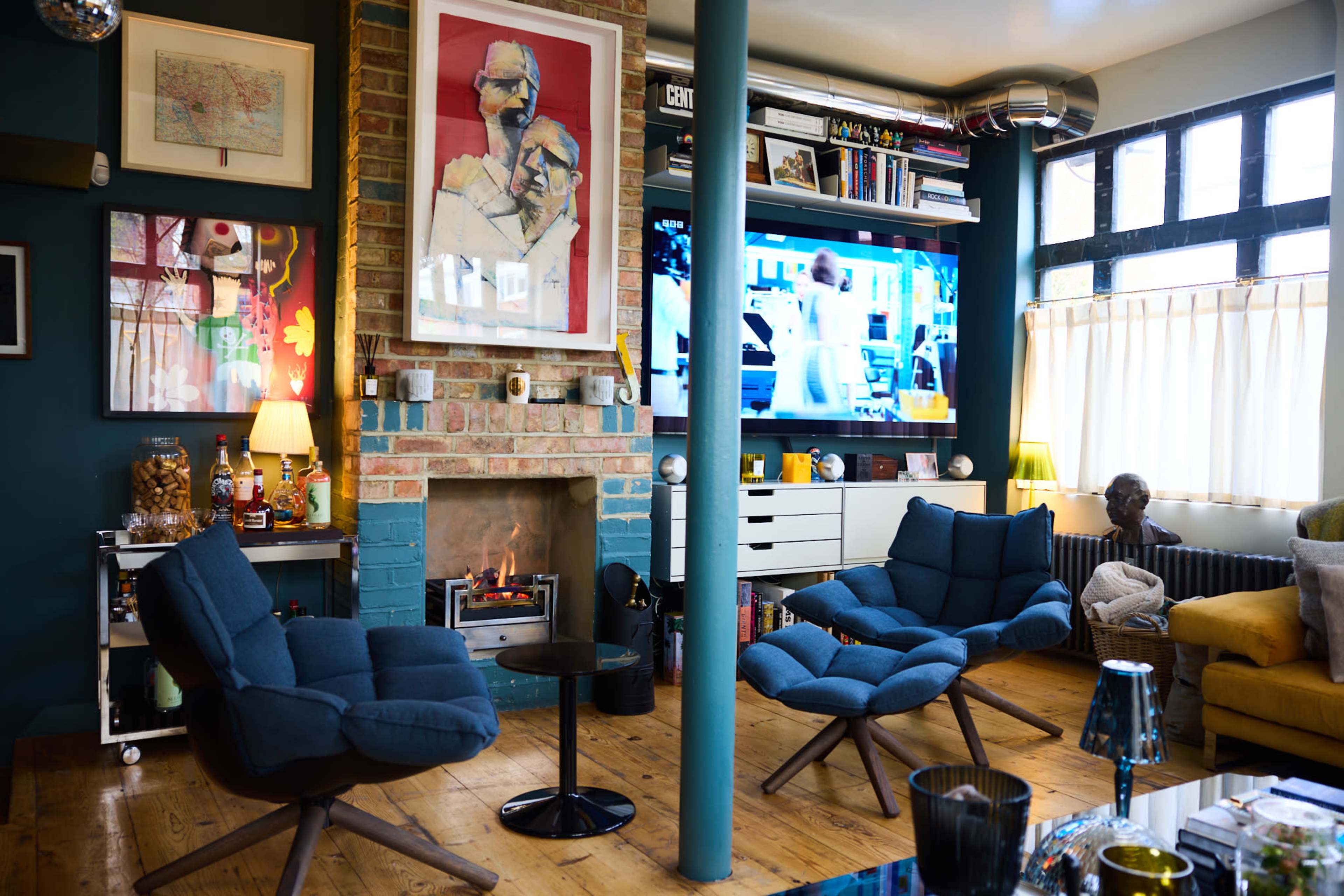 A living room with a brick fireplace, two blue armchairs, a television mounted on the wall, and various artworks and bookshelves.