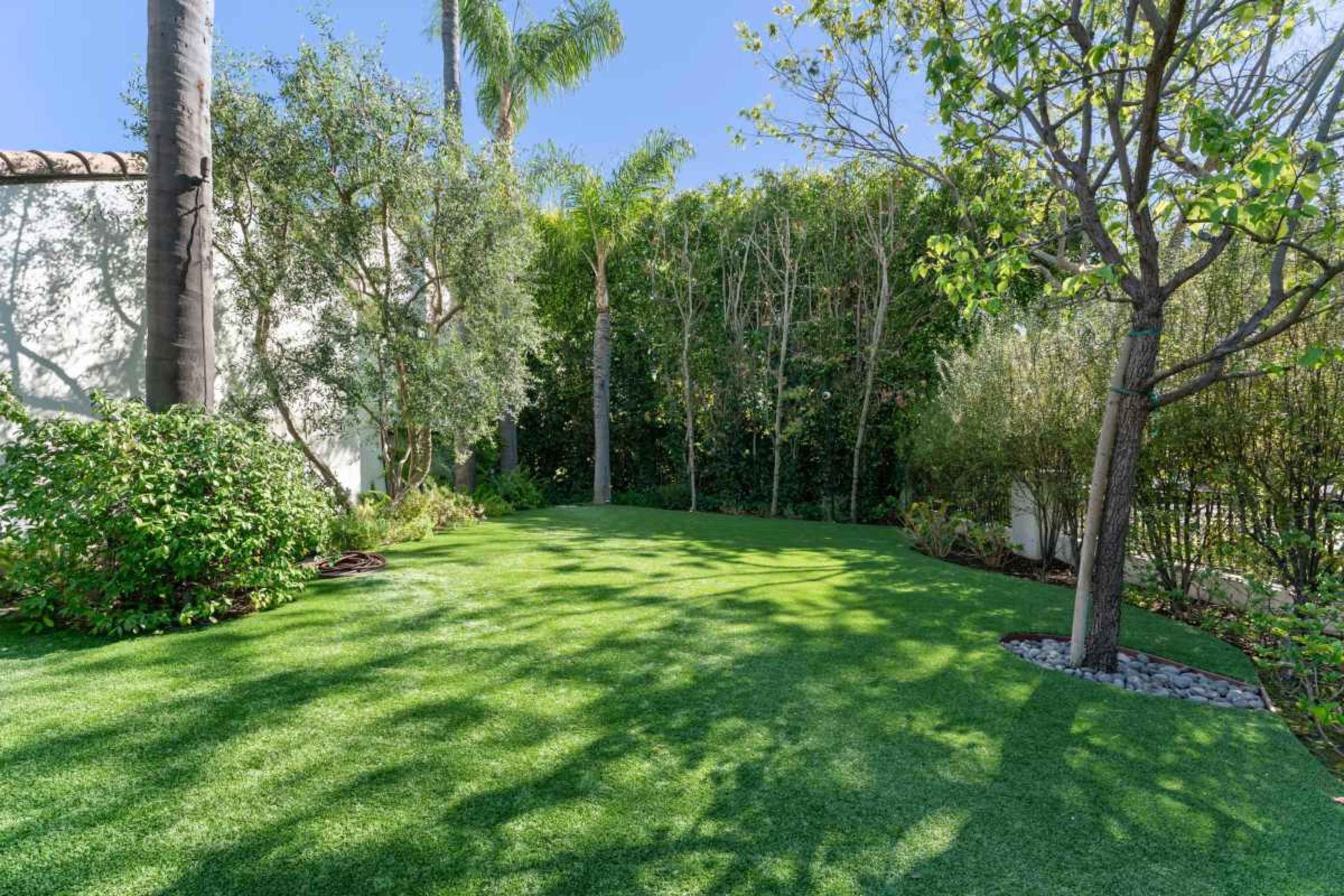 The image shows a well-maintained backyard with synthetic grass, bordered by various plants and trees.