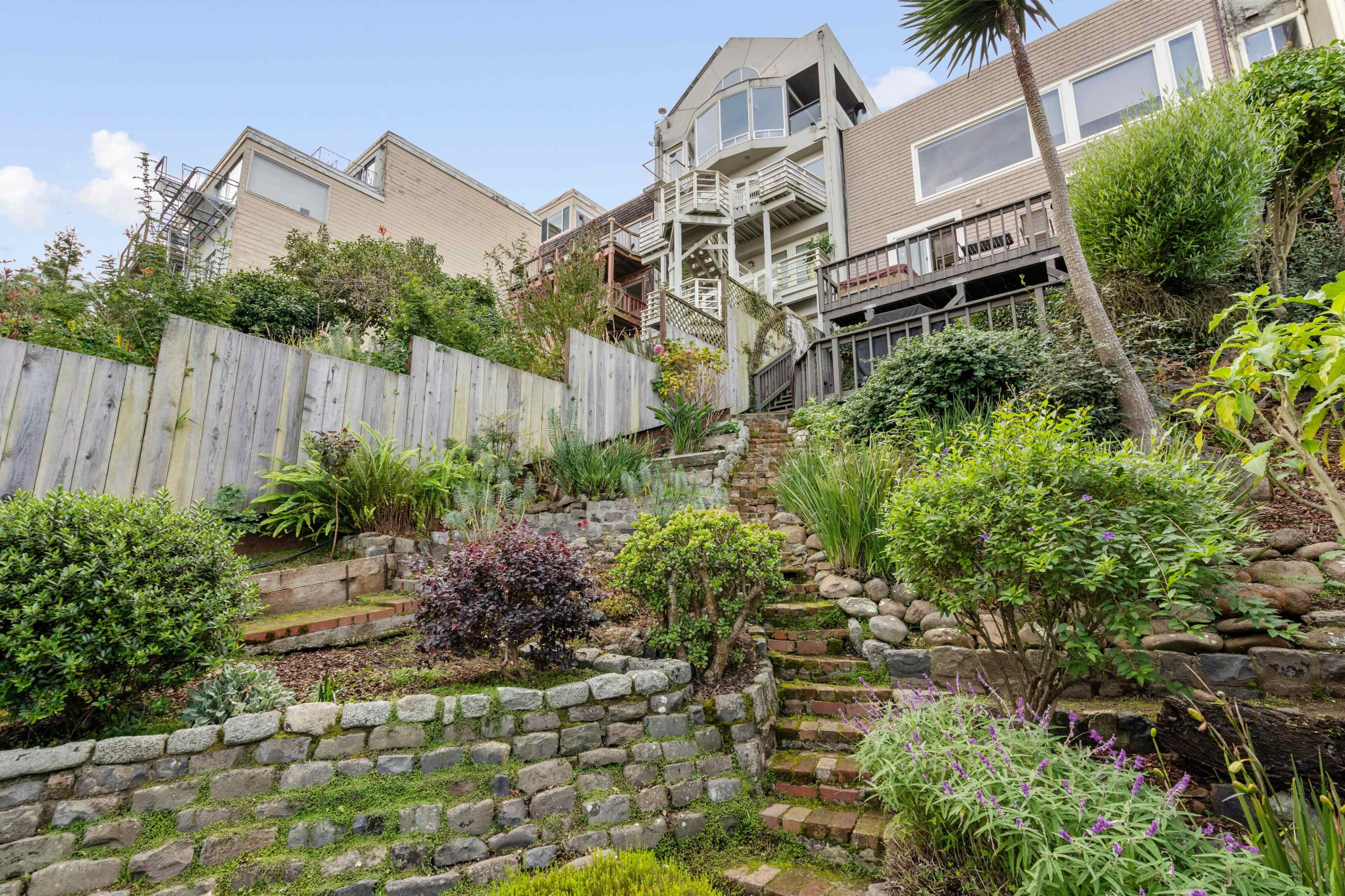 The image shows a terraced garden with stone steps leading upward to a house, surrounded by greenery and a wooden fence.