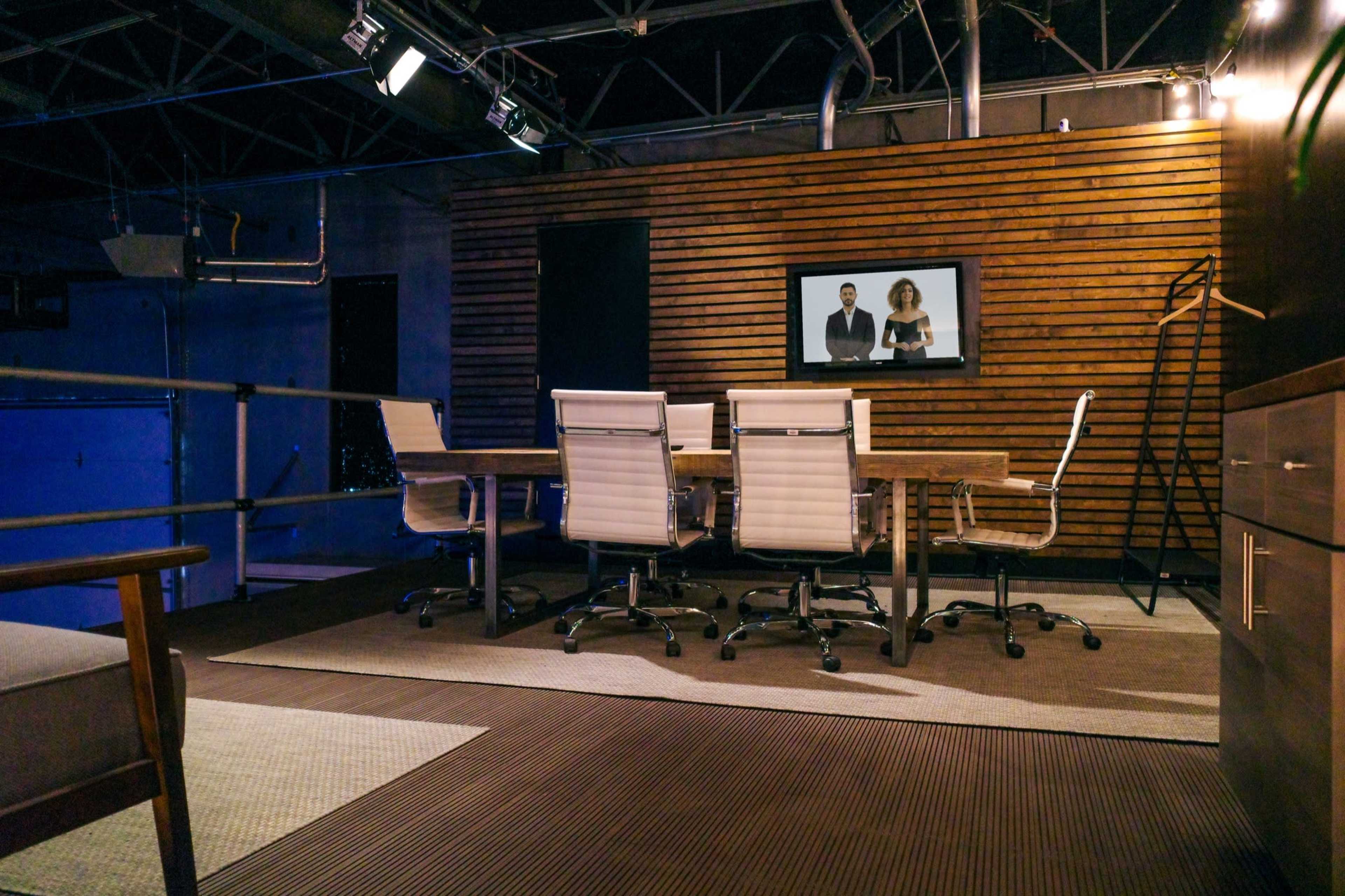 A modern conference room features a large wooden table surrounded by office chairs, with a television screen mounted on a wooden-paneled wall.