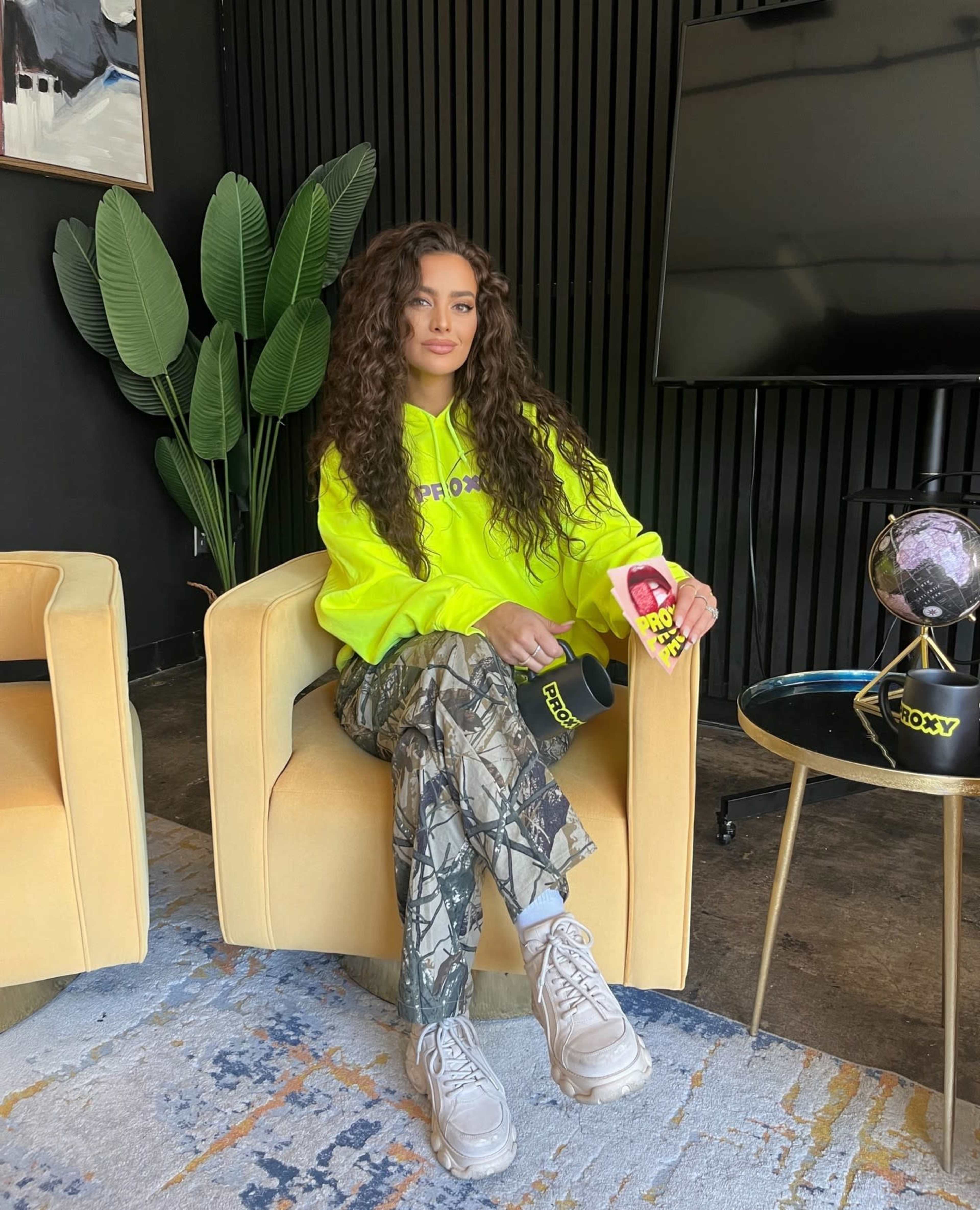 A woman sits in a yellow chair wearing a bright yellow hoodie and camo pants, holding a drink in a modern interior with a plant and a television in the background.