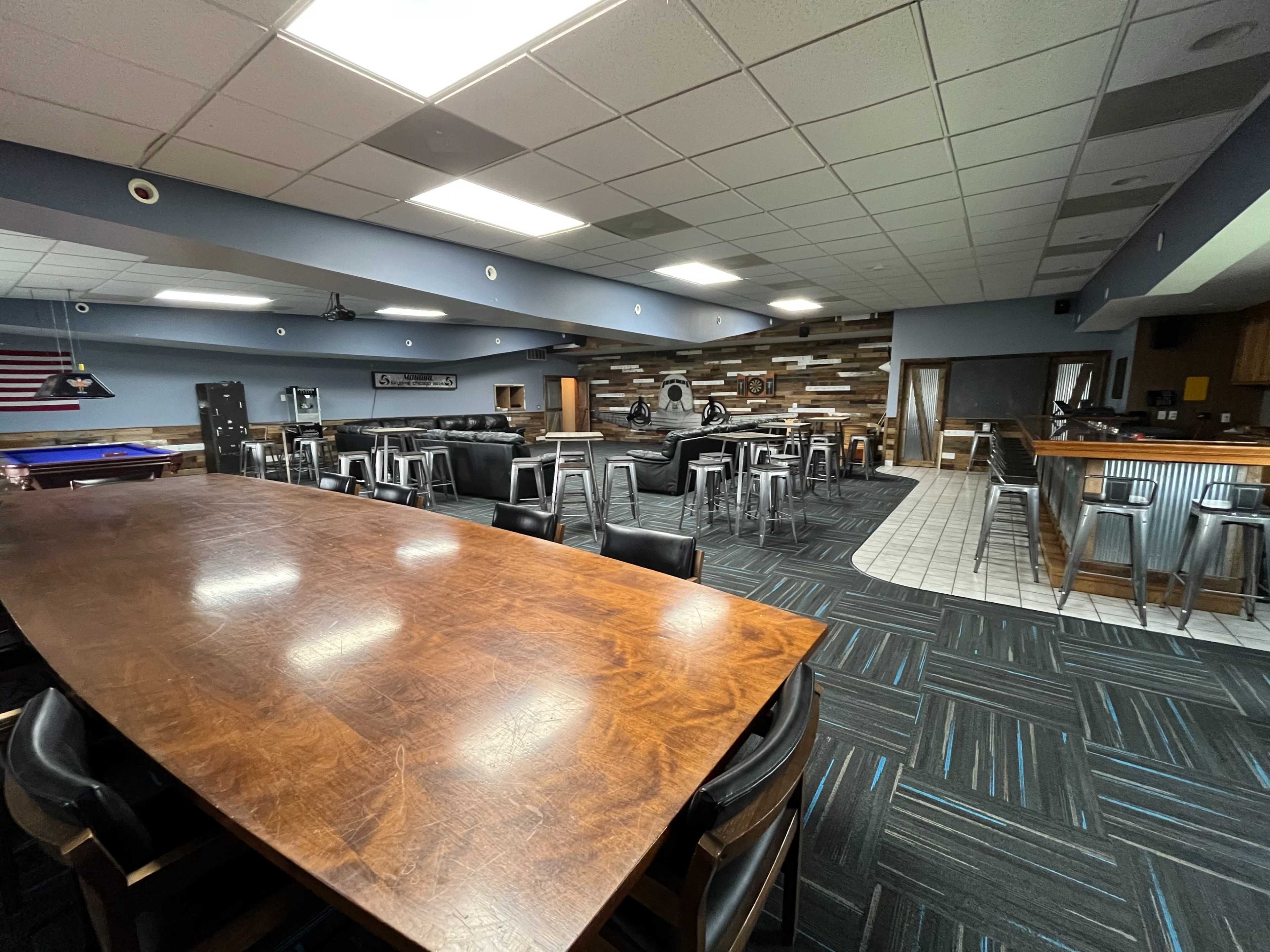 The image shows a spacious interior of a lounge or bar area featuring a long wooden table, high stools, and several seating arrangements along with a bar counter.