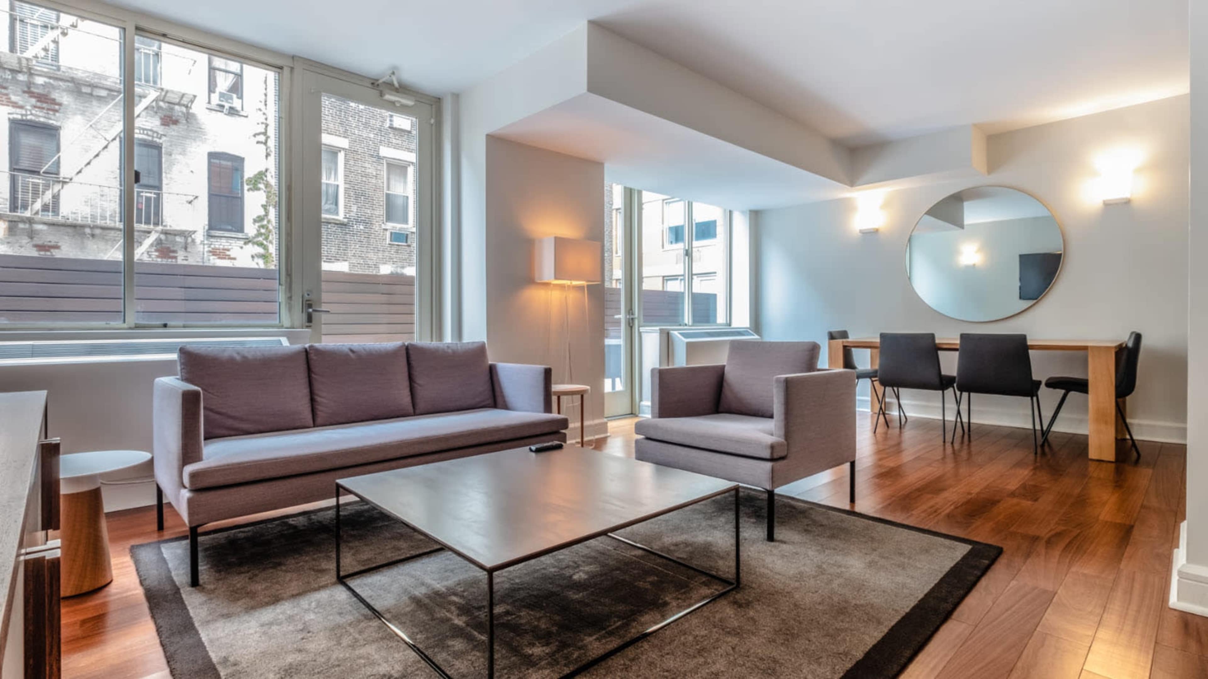 A modern living area with a gray sofa, chairs, a glass coffee table, and a dining table in a well-lit space with large windows.