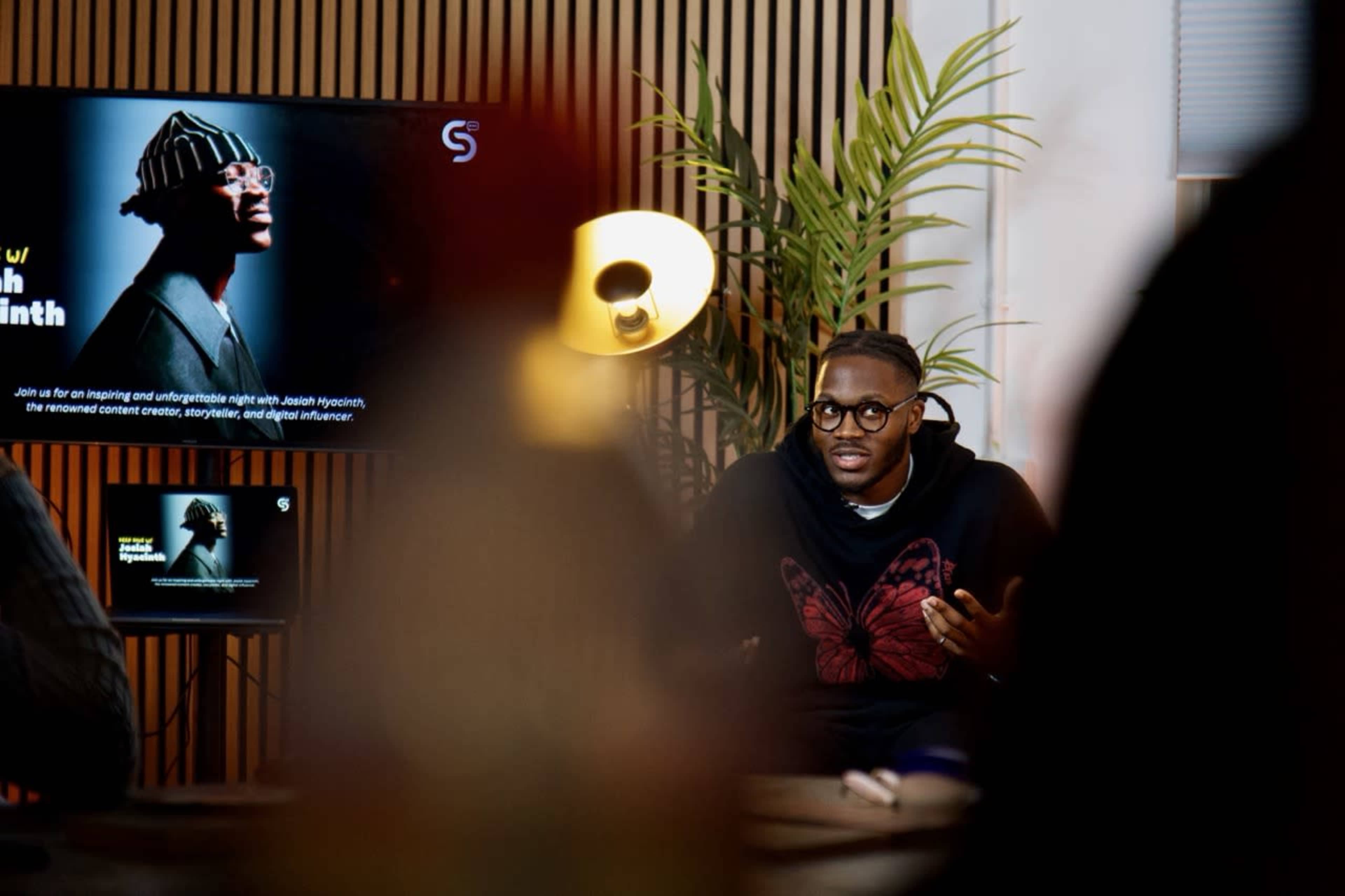 A man with glasses and a butterfly graphic on his shirt speaks during a presentation in a modern office setting, with screens displaying promotional materials in the background.