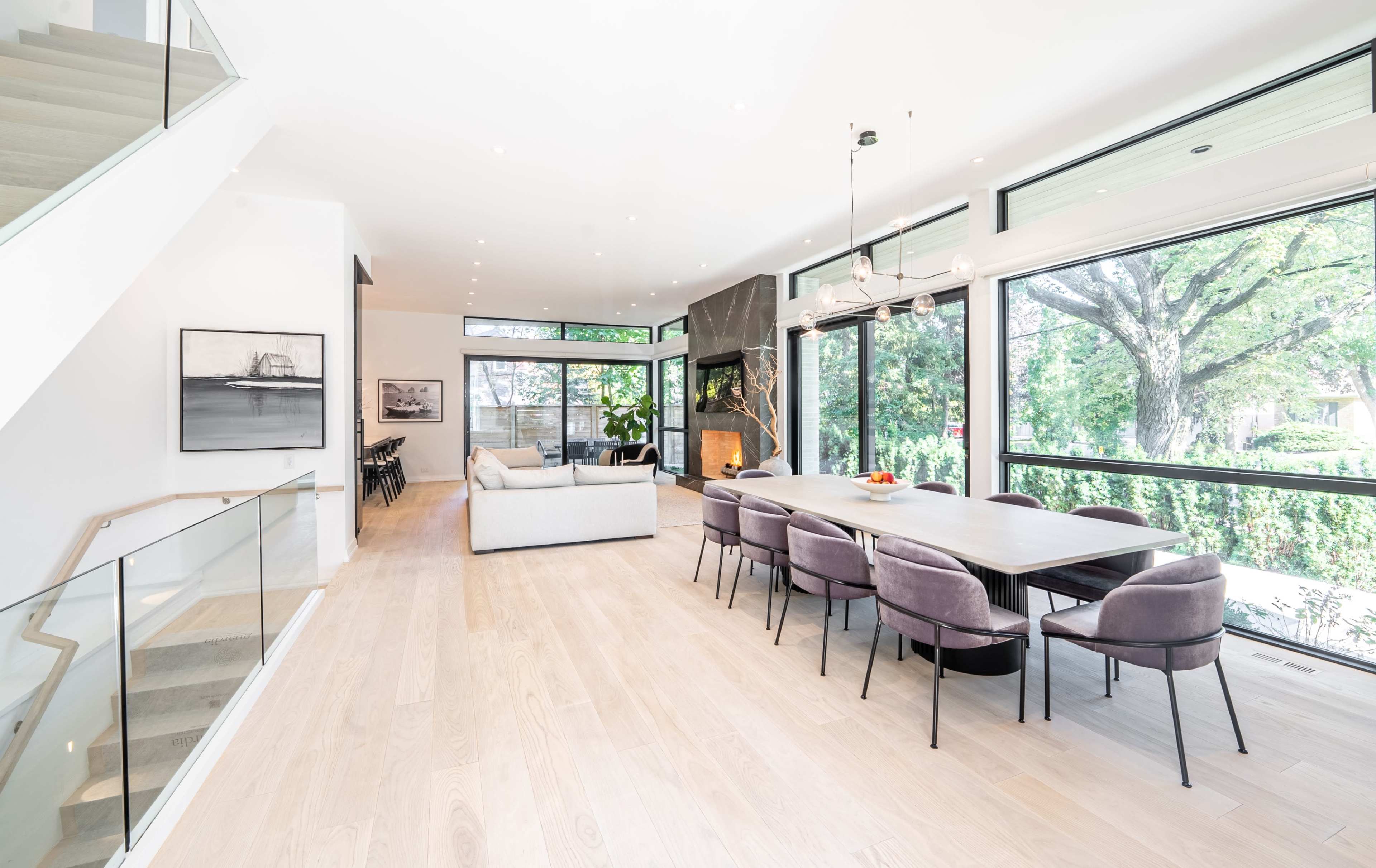 The image shows a modern open-concept living and dining area with large windows, a fireplace, and a dining table surrounded by plush chairs.