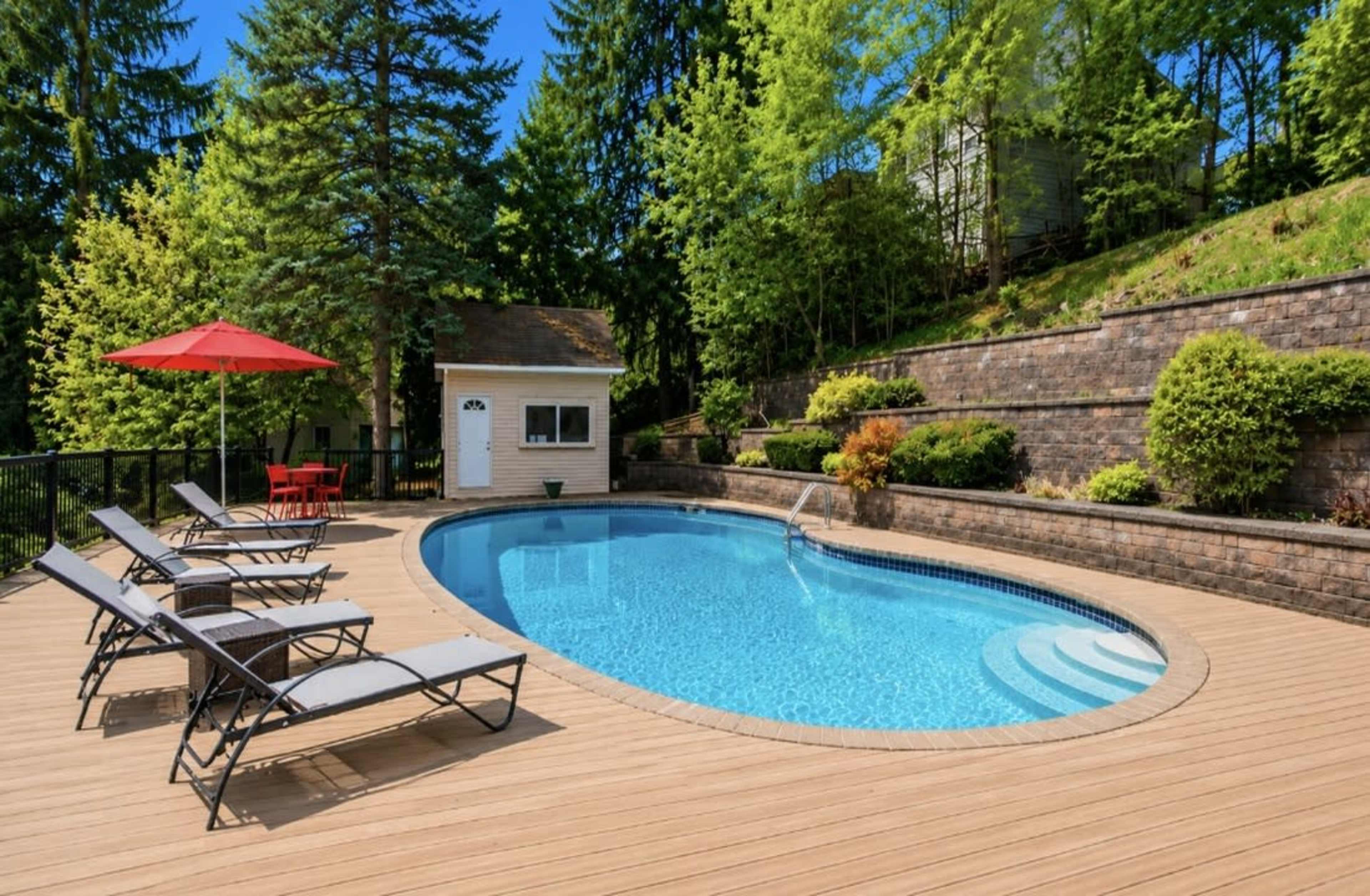 The image shows a rectangular swimming pool surrounded by wooden decking, lounge chairs, and a red umbrella, with greenery and a small building in the background.
