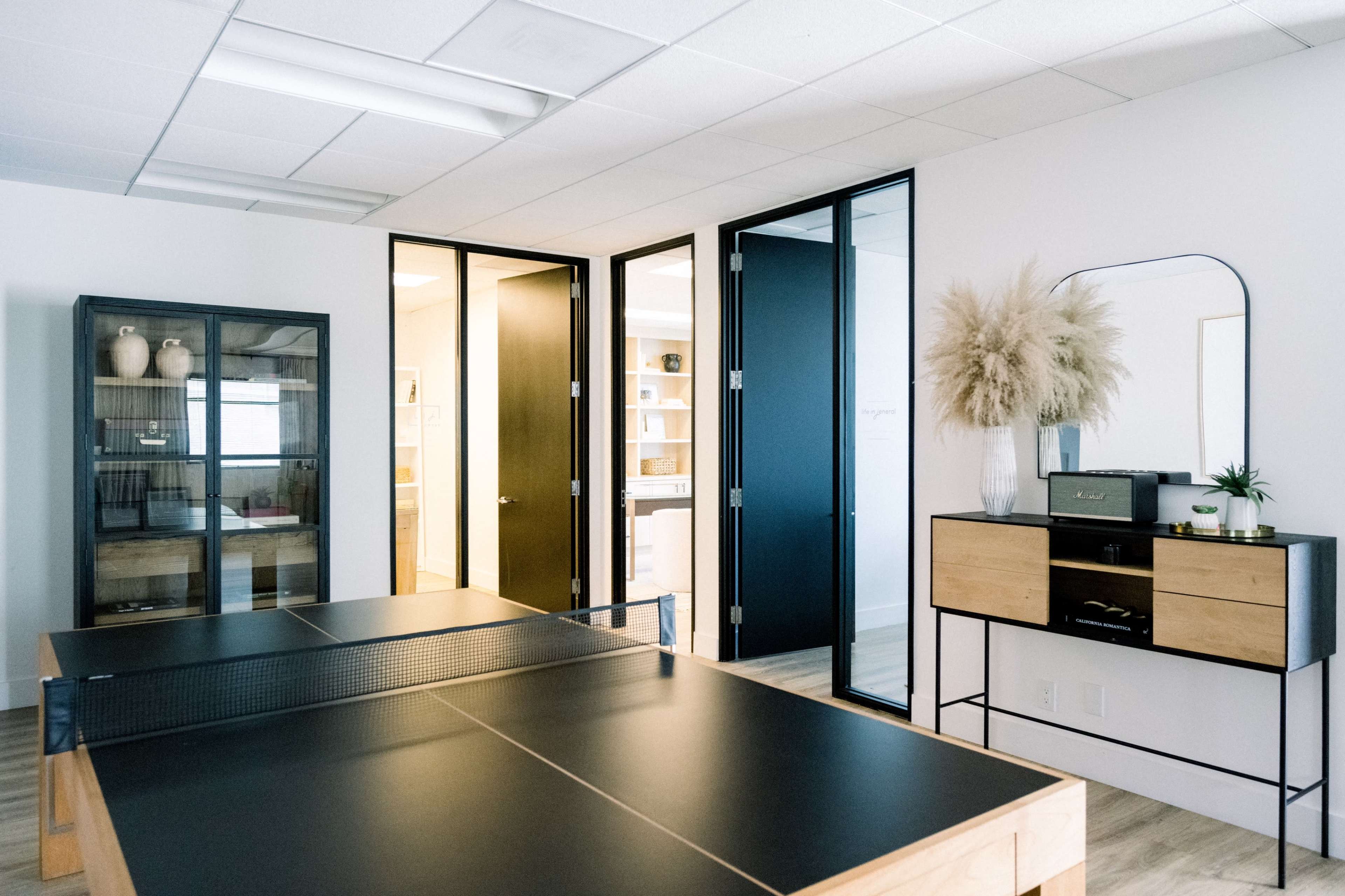 A modern office space features a ping pong table, black and wood furniture, and several closed doors leading to other areas.