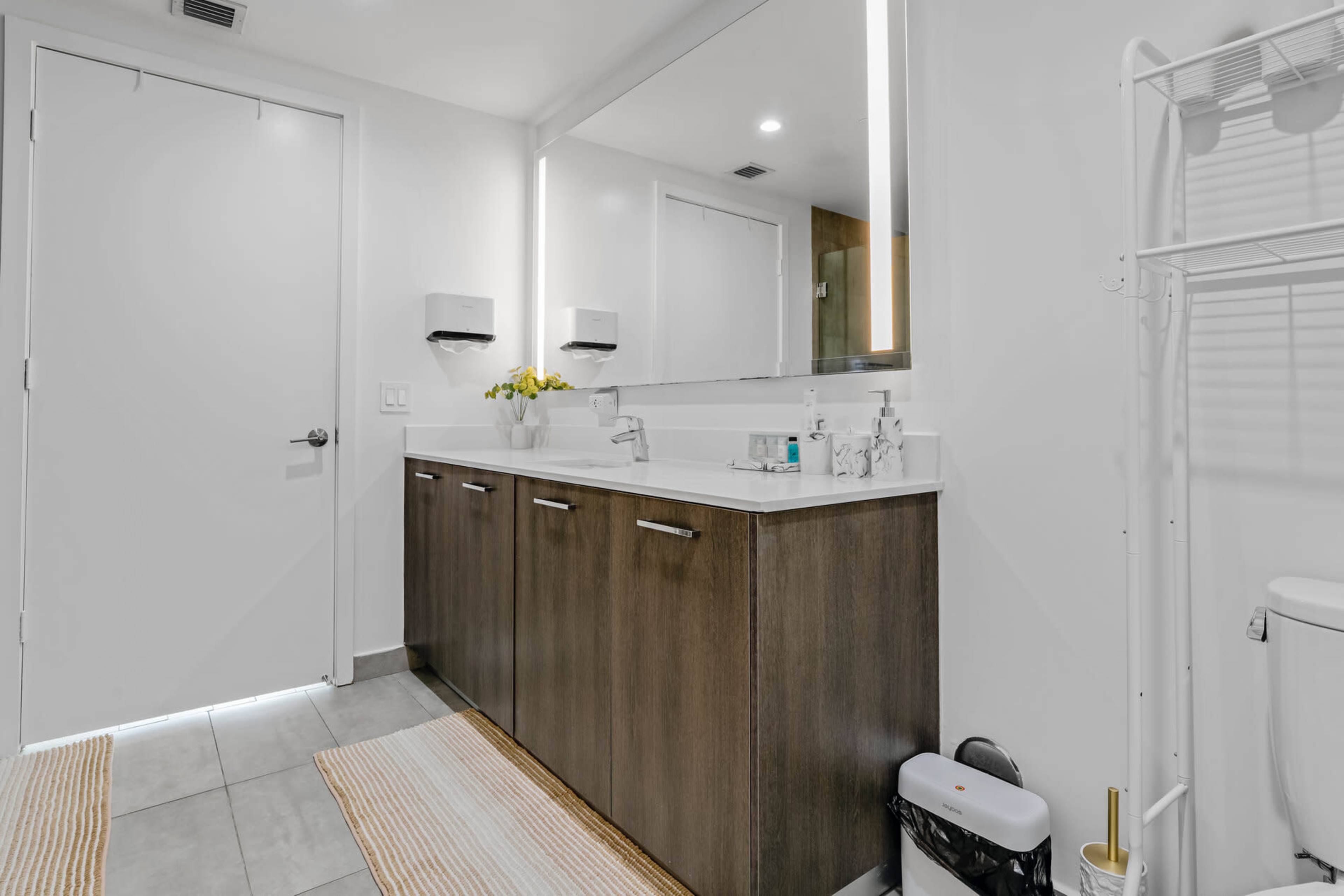 The image shows a modern bathroom featuring a wooden vanity with a white countertop, a large mirror, and a clean, minimalist design.