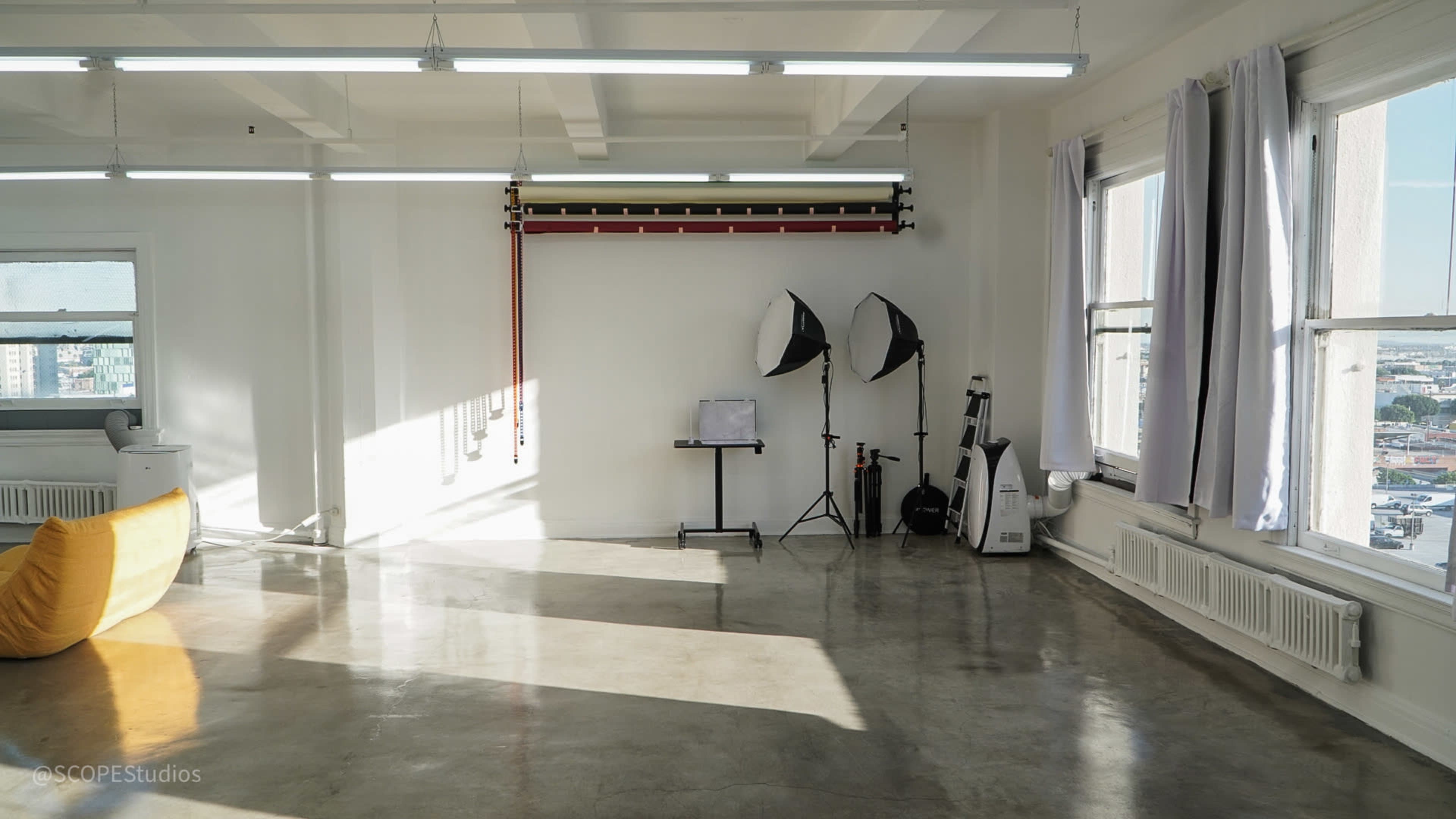 The image shows a bright, minimalist studio space with polished concrete floors, large windows, and basic equipment for photography.