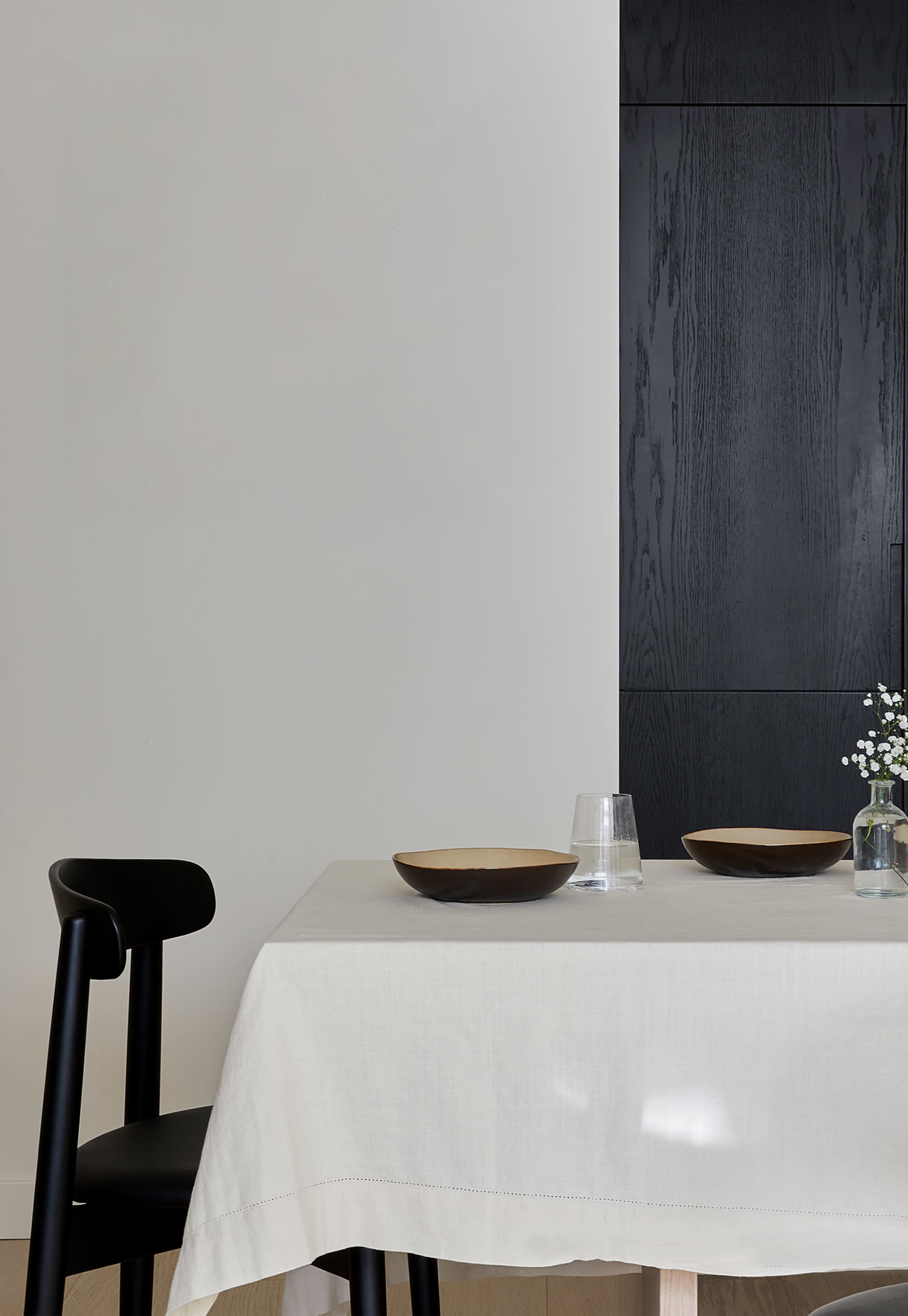 A minimalist dining table is set with two bowls and a glass, accompanied by a black chair and a small vase of flowers against a light-colored wall.