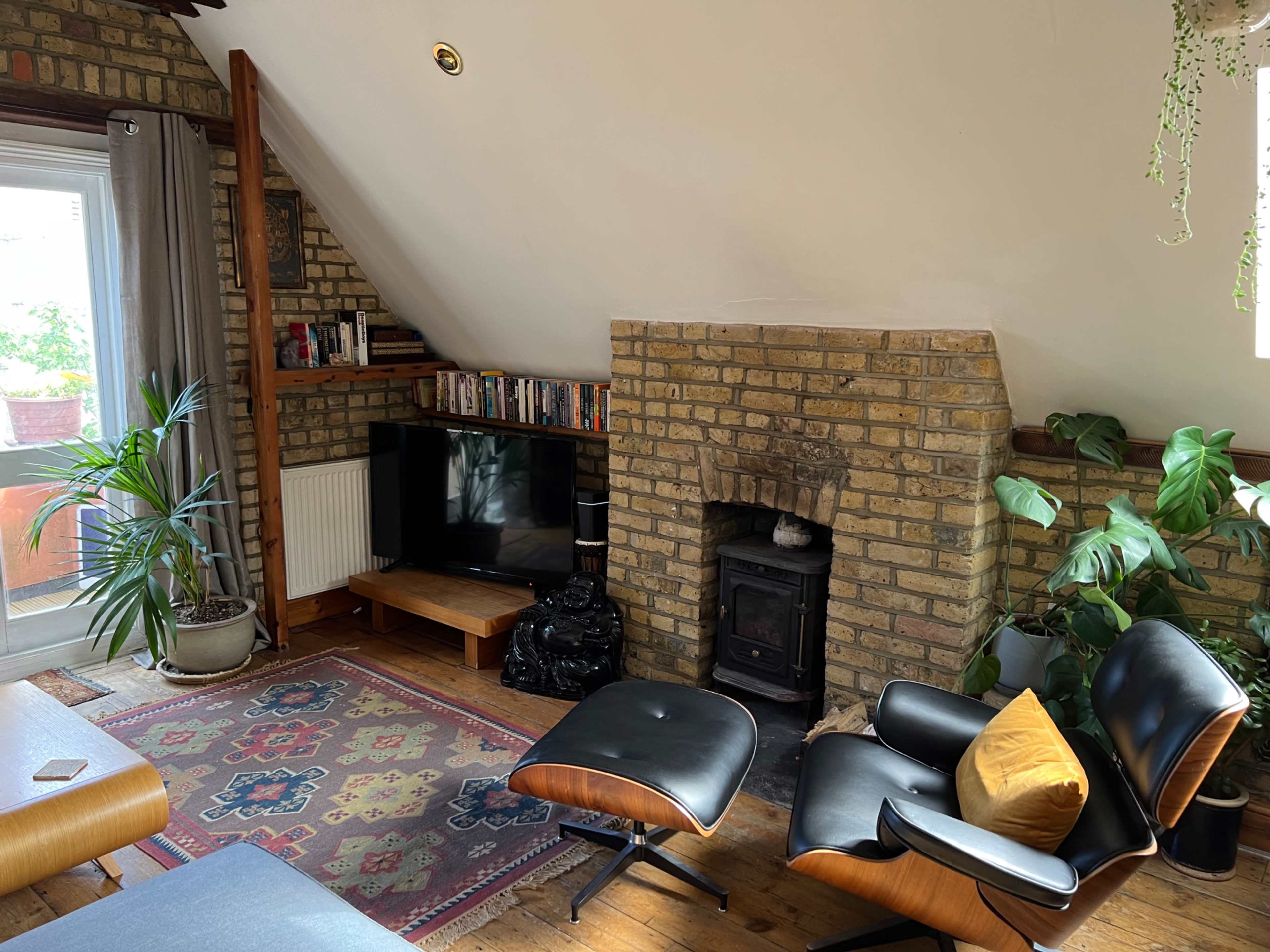 The image shows a cozy living room with a brick fireplace, a television on a stand, a large window, and comfortable seating with plants around.