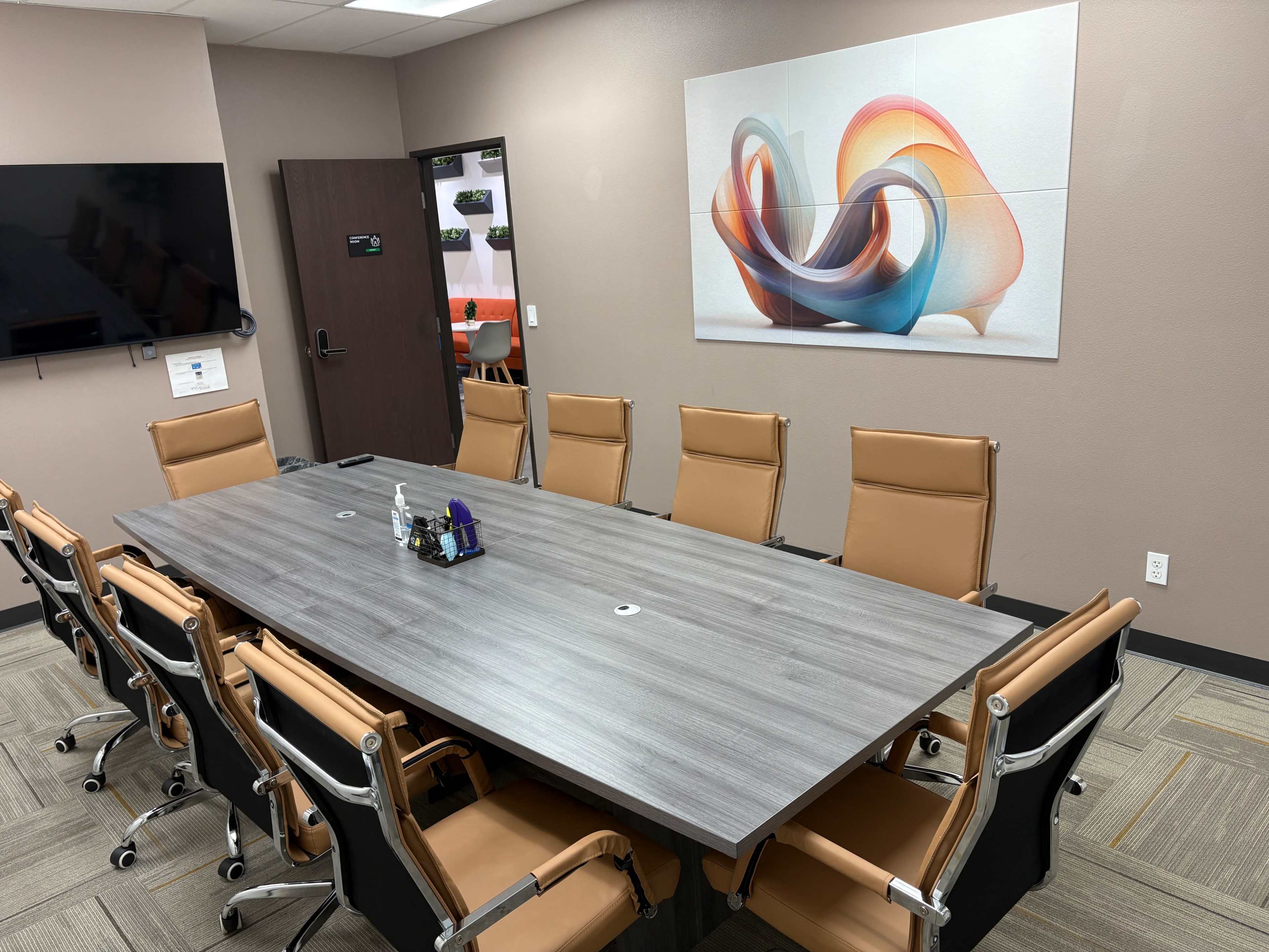 The image shows a conference room with a large rectangular table surrounded by brown leather chairs, and a colorful abstract artwork mounted on the wall.
