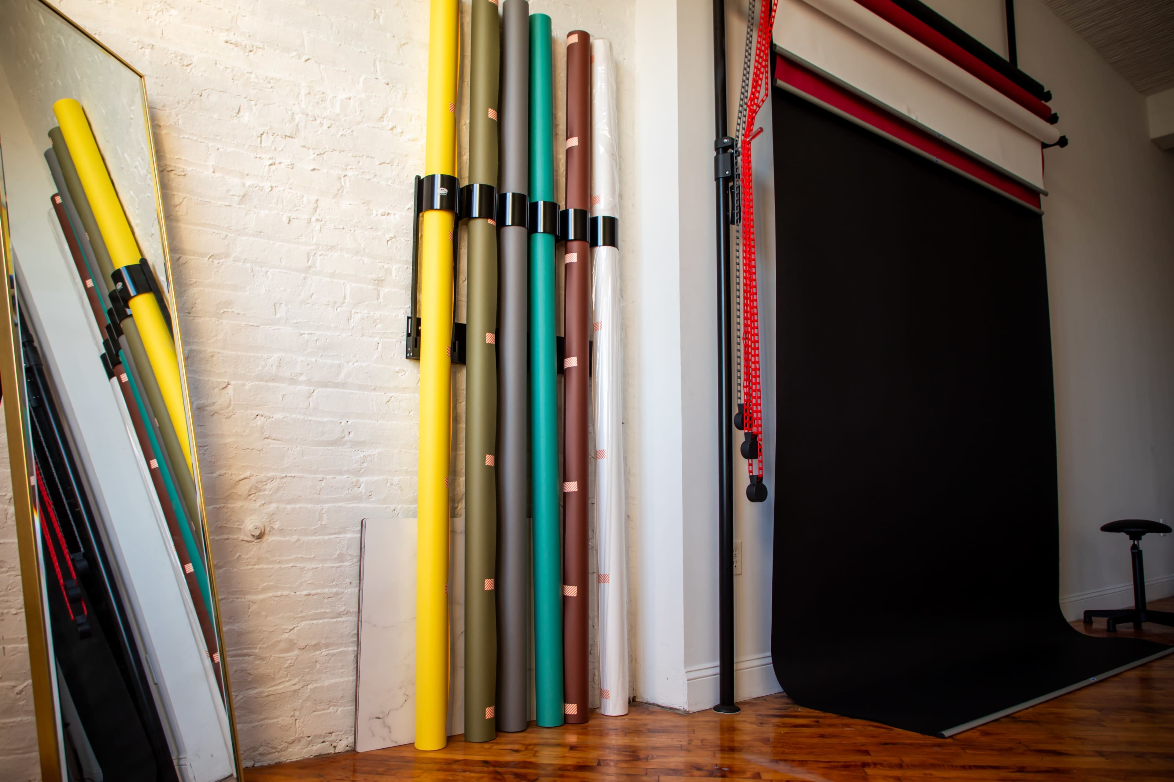 The image shows a collection of rolled photo backdrop materials in various colors stacked against a white brick wall, alongside a large black backdrop hanging on a frame.