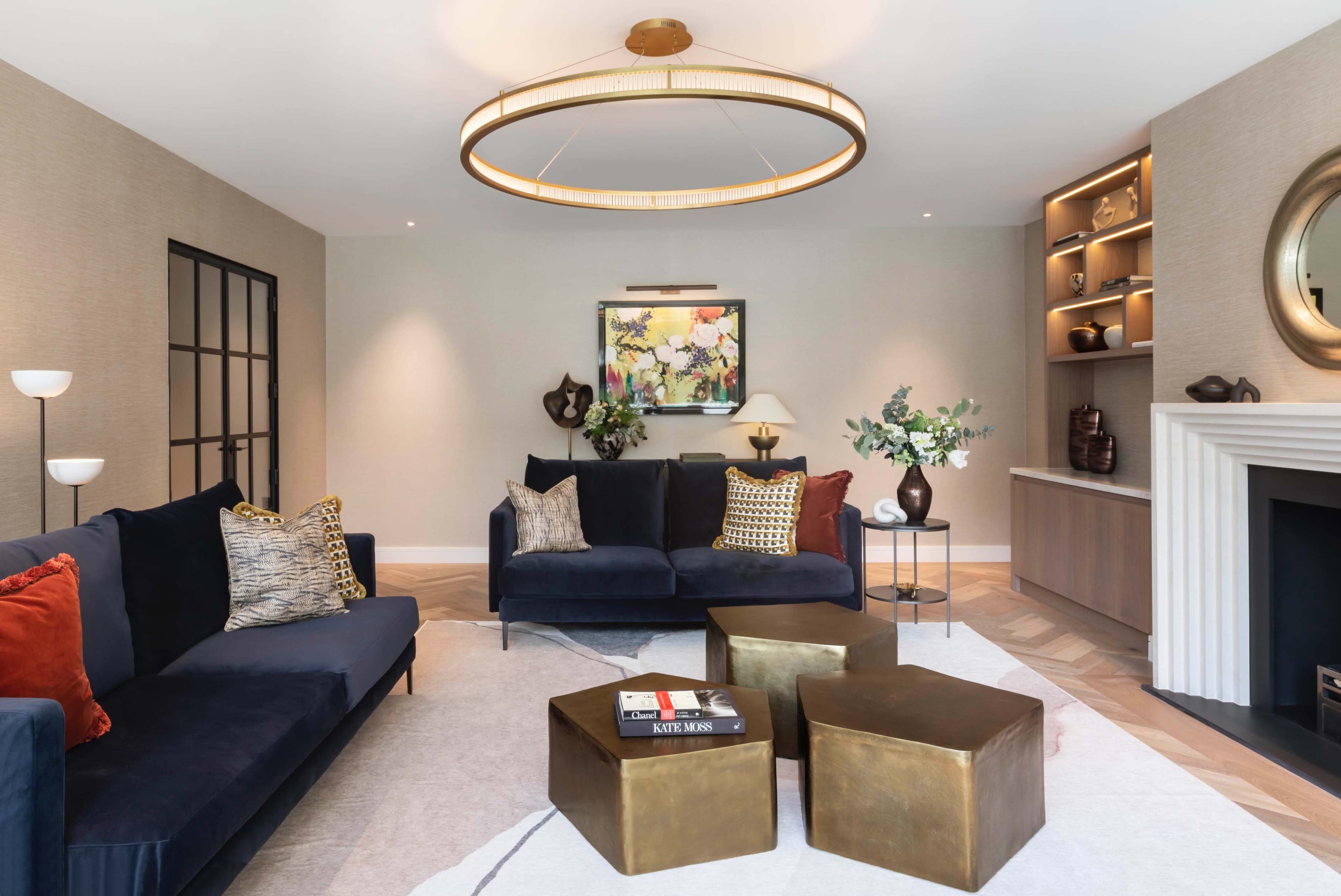 A modern living room features dark blue sofas, brass accent tables, a large circular light fixture, and decorative art on the walls.