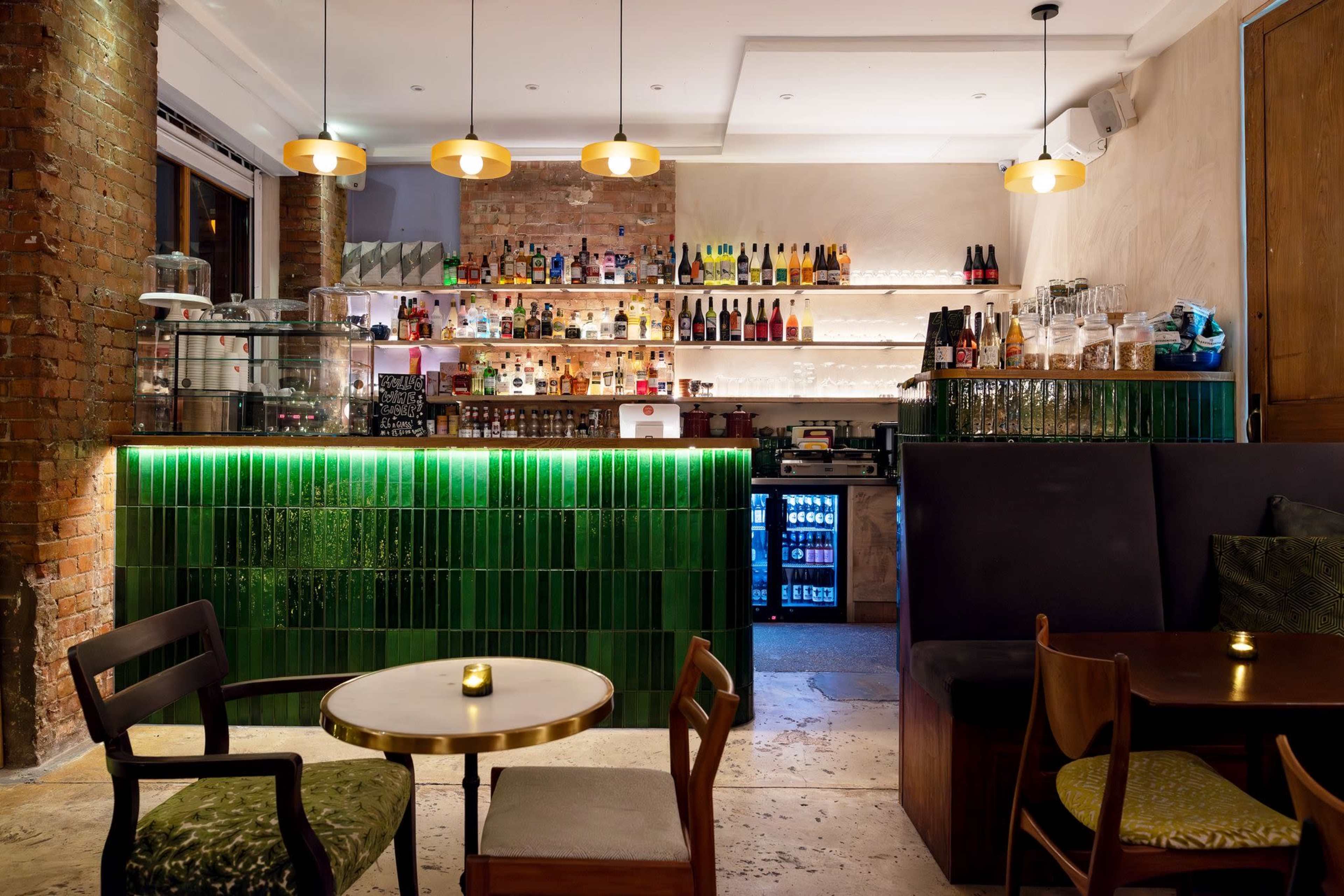 The image shows an elegantly designed bar area with a green tile counter, shelves filled with various bottles, and soft lighting, accompanied by wooden tables and chairs.