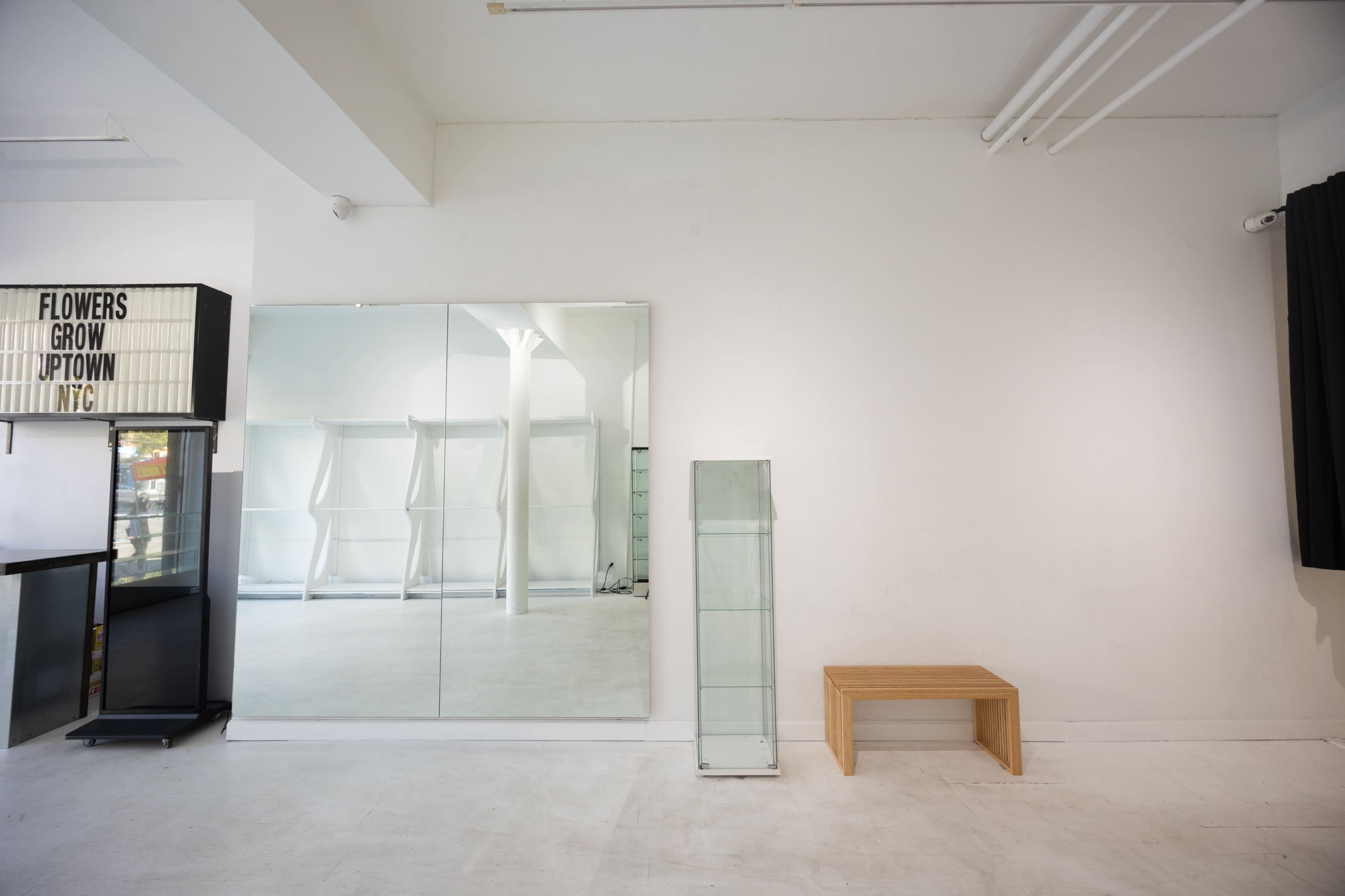 The image shows a minimalist interior space featuring a large mirror, a glass display case, and a wooden bench against white walls.