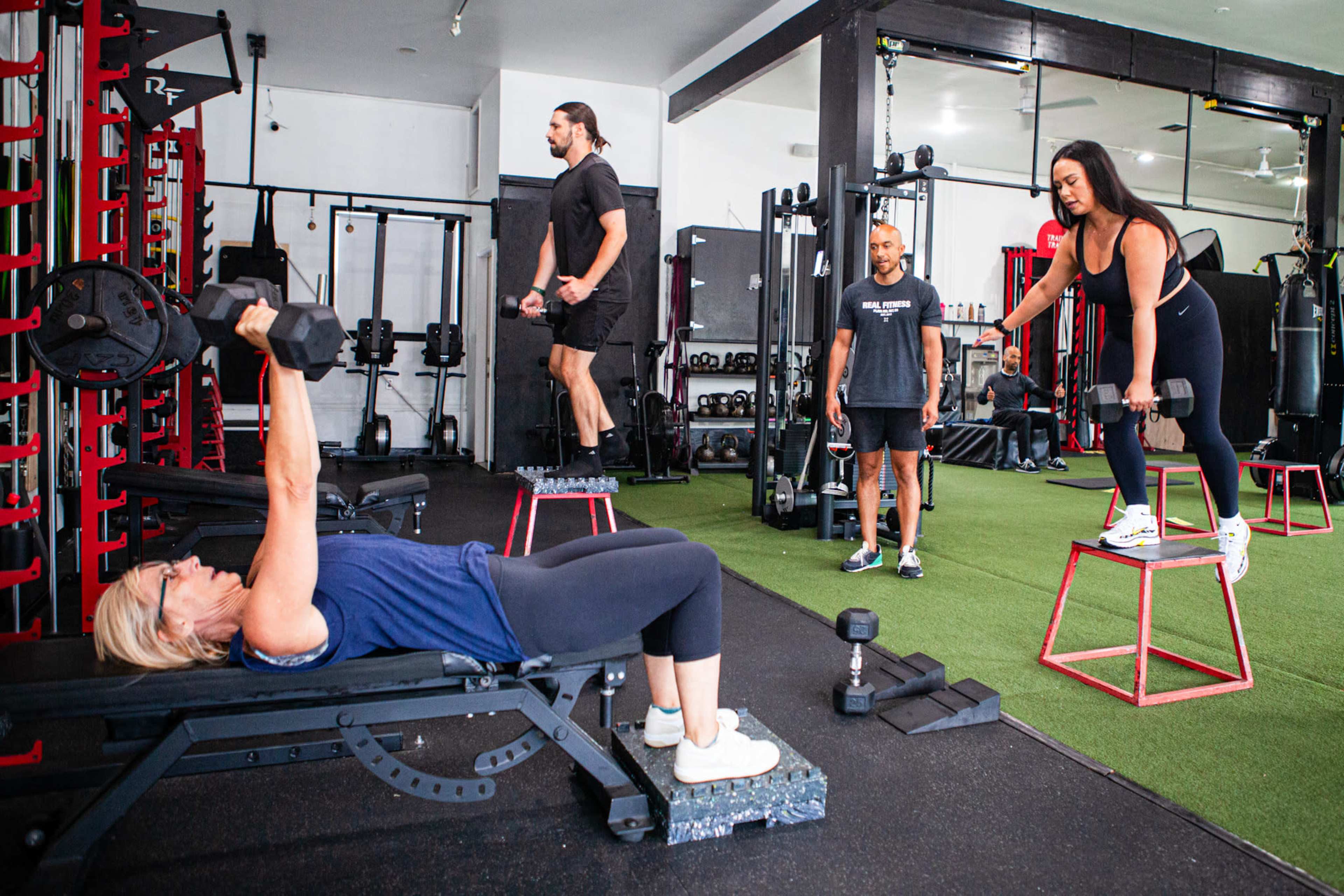 A group of four people engages in various weightlifting exercises in a gym setting.