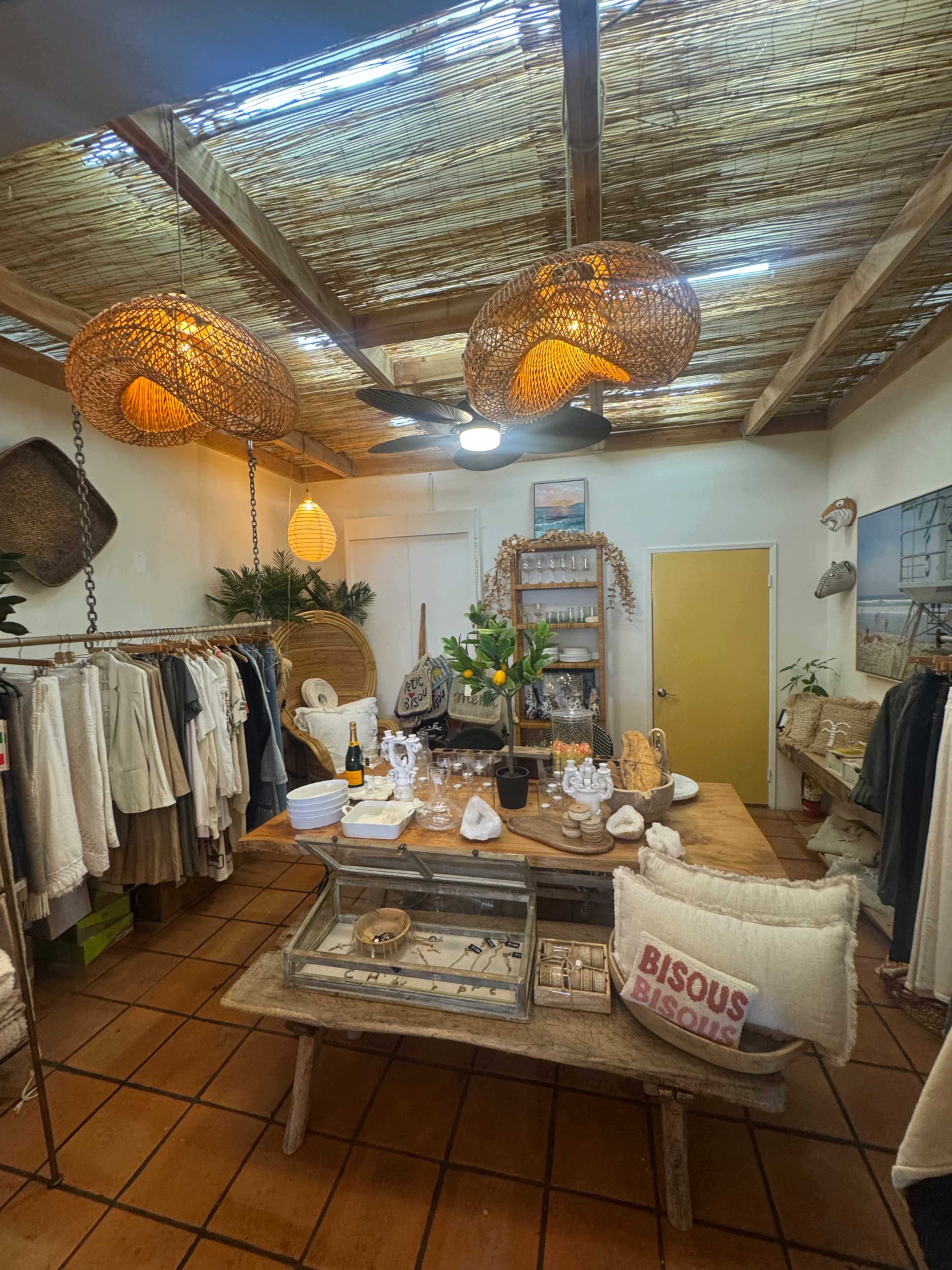The image shows a cozy shop interior with wooden furniture, hanging light fixtures, and clothing on racks along the walls.