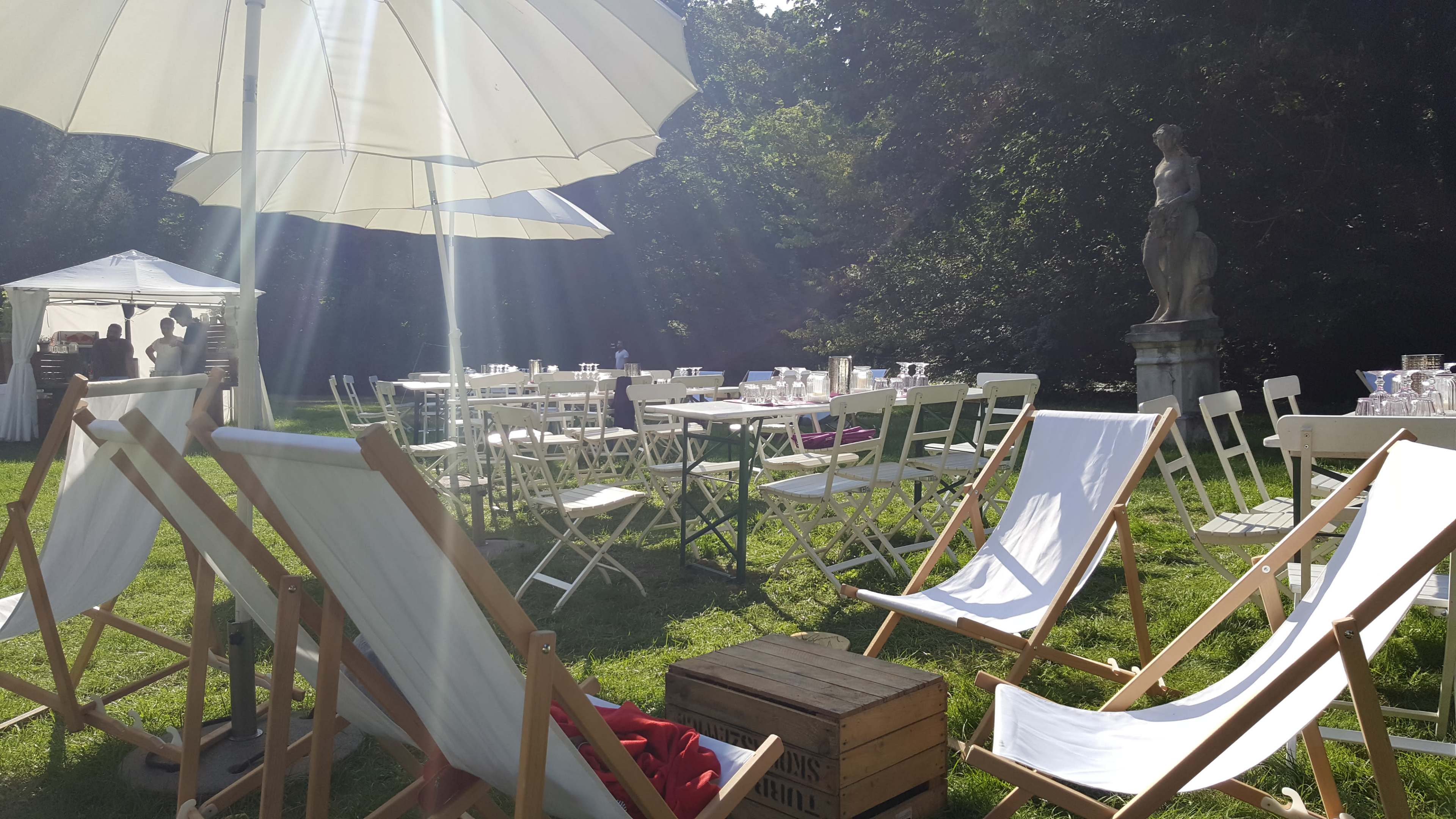 The scene depicts an outdoor event area featuring rows of white tables and chairs, a large umbrella, and lounge chairs arranged on green grass.