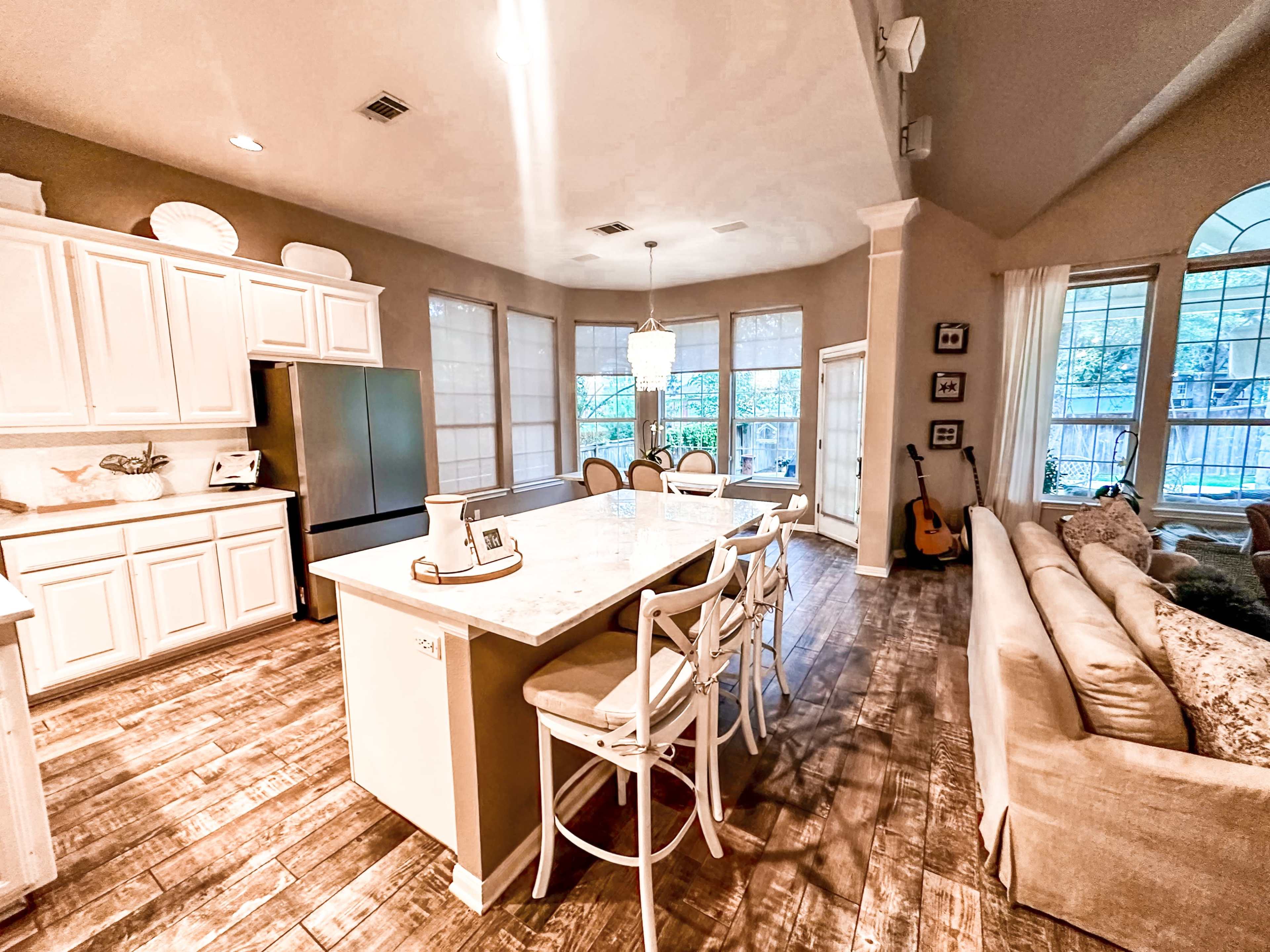 The image shows a modern kitchen and living area with light-colored cabinetry, a large island with bar stools, and a seating area featuring a sofa, all set against a backdrop of large windows.