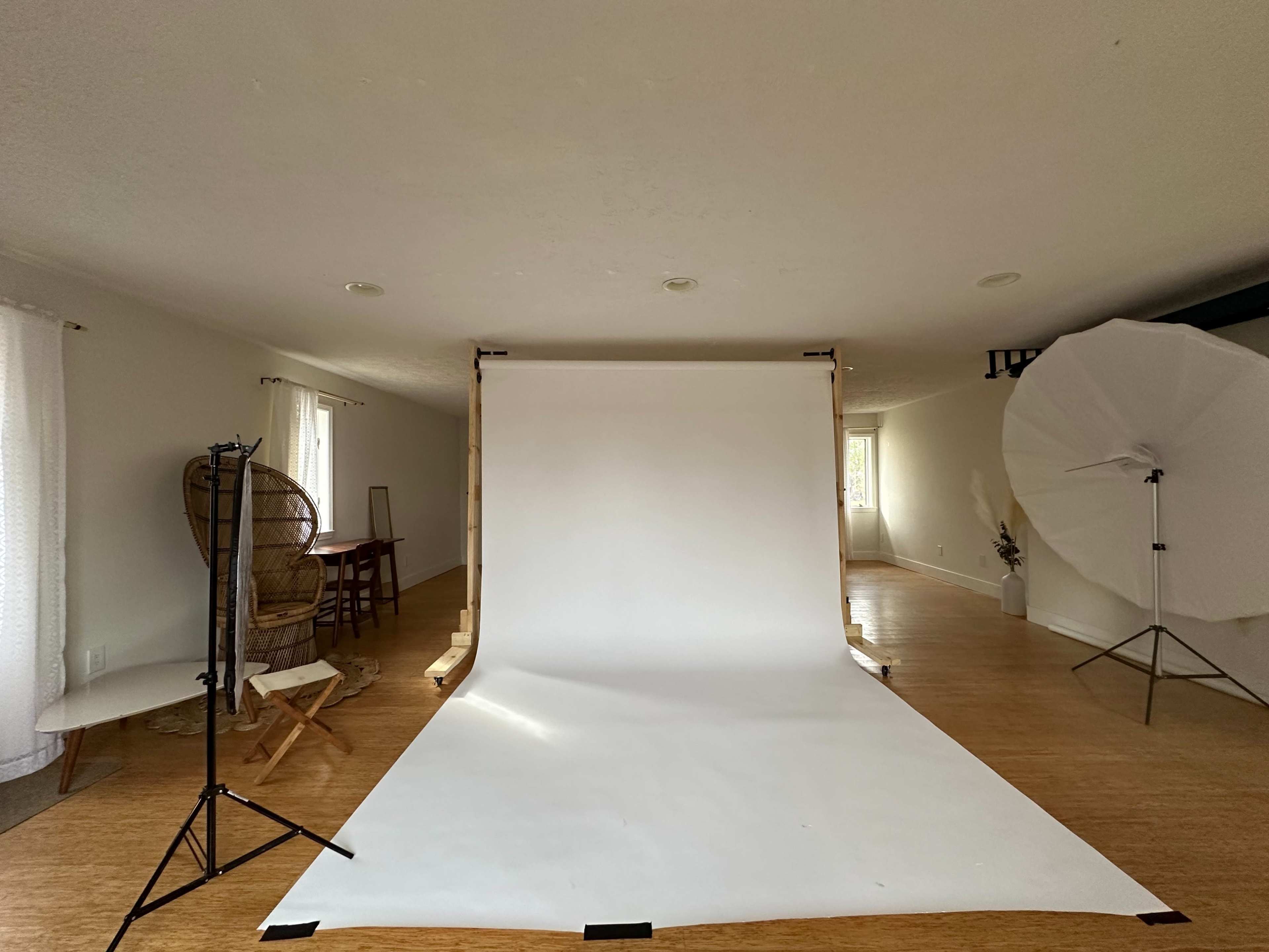 A photography studio setup featuring a white backdrop, a lighting umbrella, and a chair in a bright room.