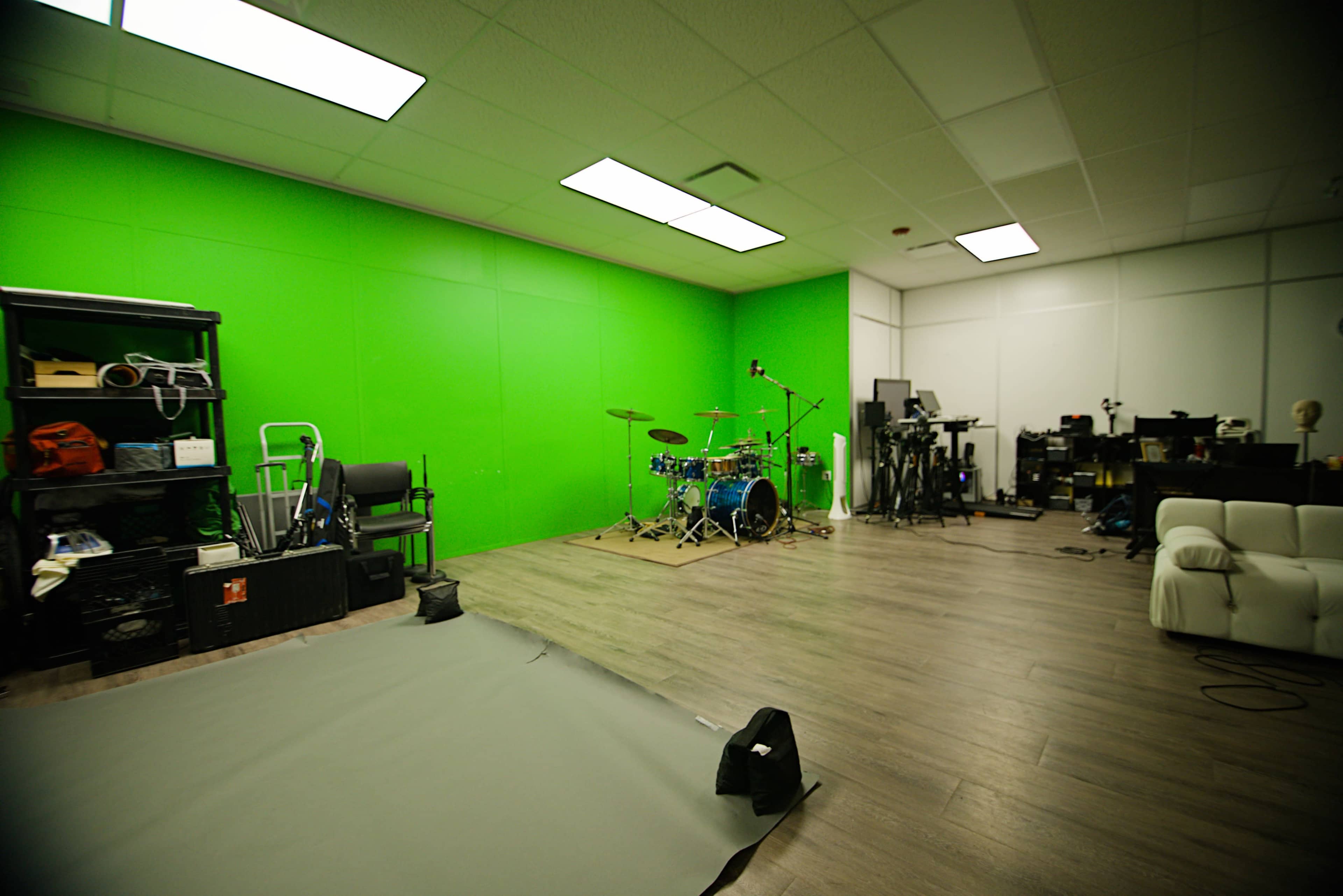 The image shows a spacious studio with a green screen, a drum set, and various music and recording equipment.
