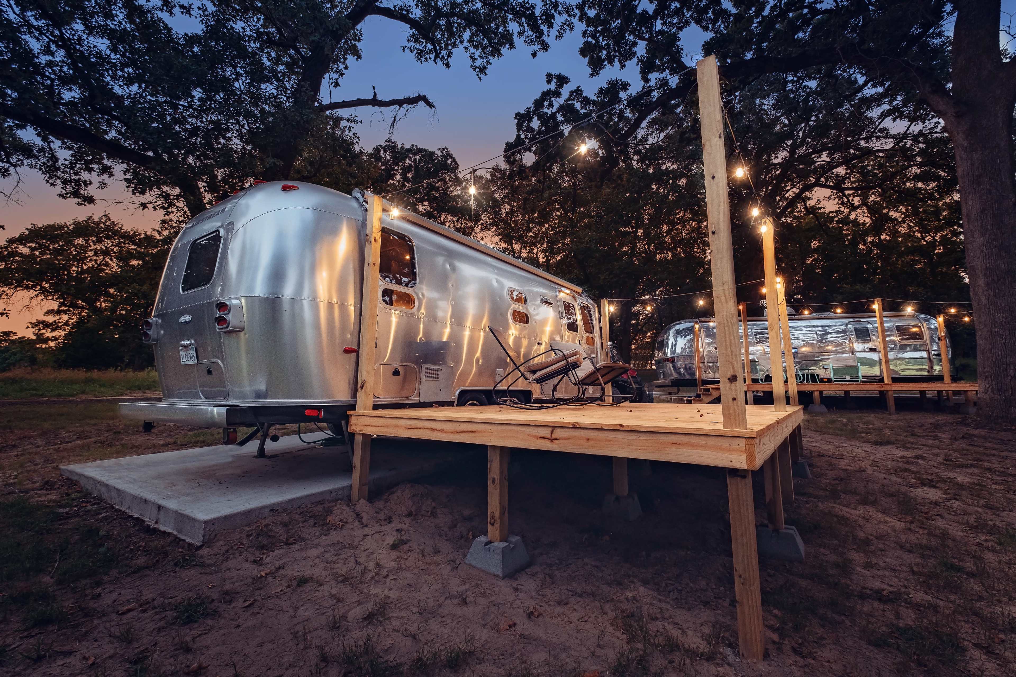 A silver Airstream trailer is parked on a wooden platform surrounded by trees, with string lights illuminating the scene during twilight.